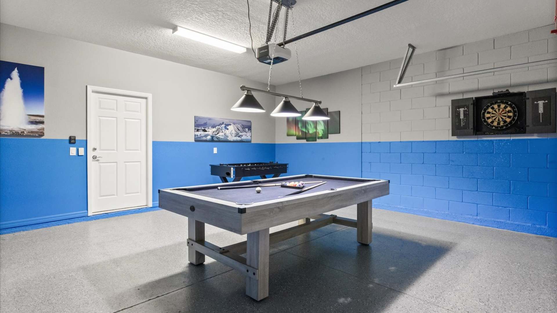 Garage converted into a game room with a pool table as the centerpiece, a foosball table, and a dartboard on the wall. The walls are painted gray and blue, and the floor is a speckled gray epoxy.