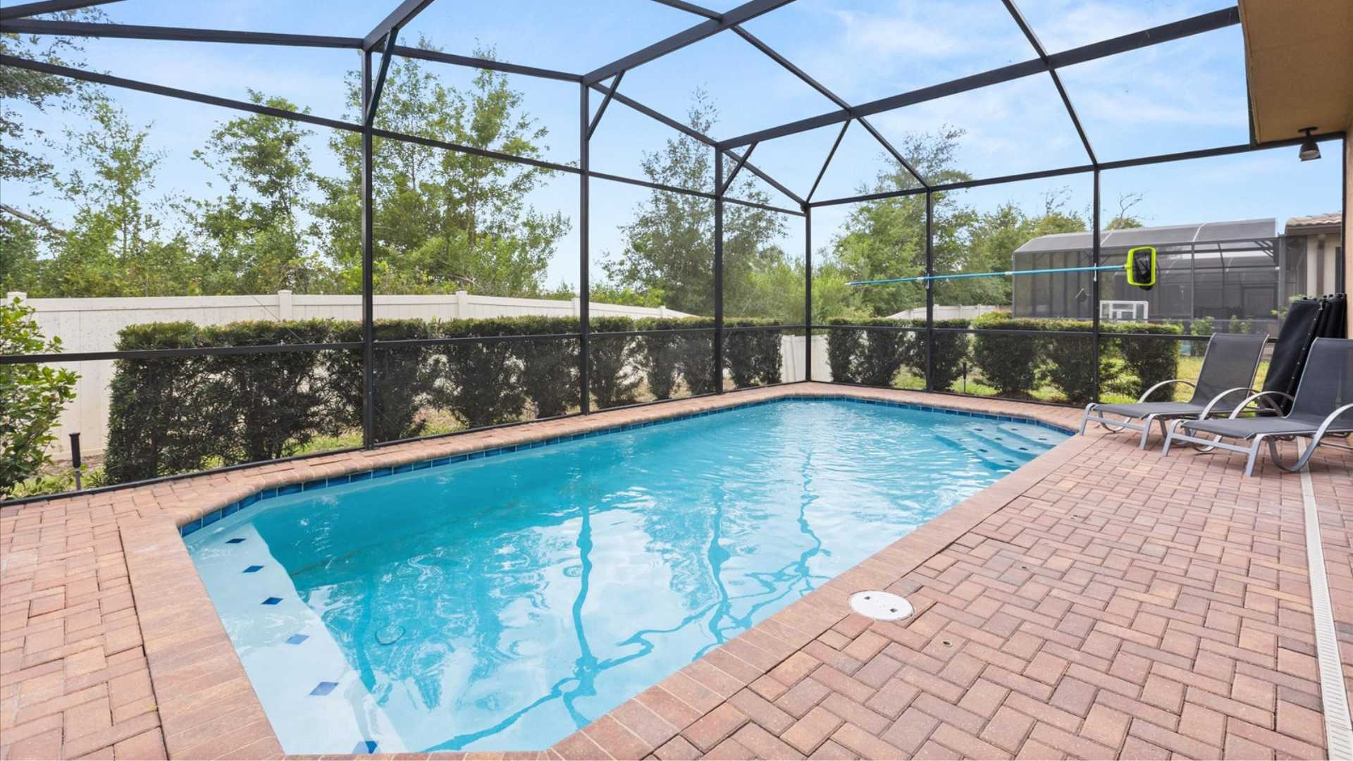 Rectangular private swimming pool with blue tile accents, surrounded by a red brick paver deck and enclosed by a black screen cage. Lush green landscaping and a fence are visible beyond the cage.