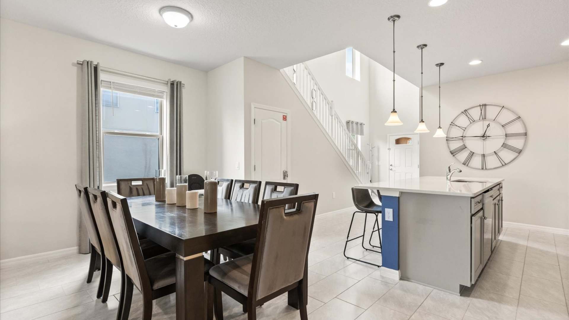 Spacious dining area with a large dark wood table and upholstered chairs, adjacent to a kitchen island with pendant lights, and a grand staircase leading to the upper floor.
