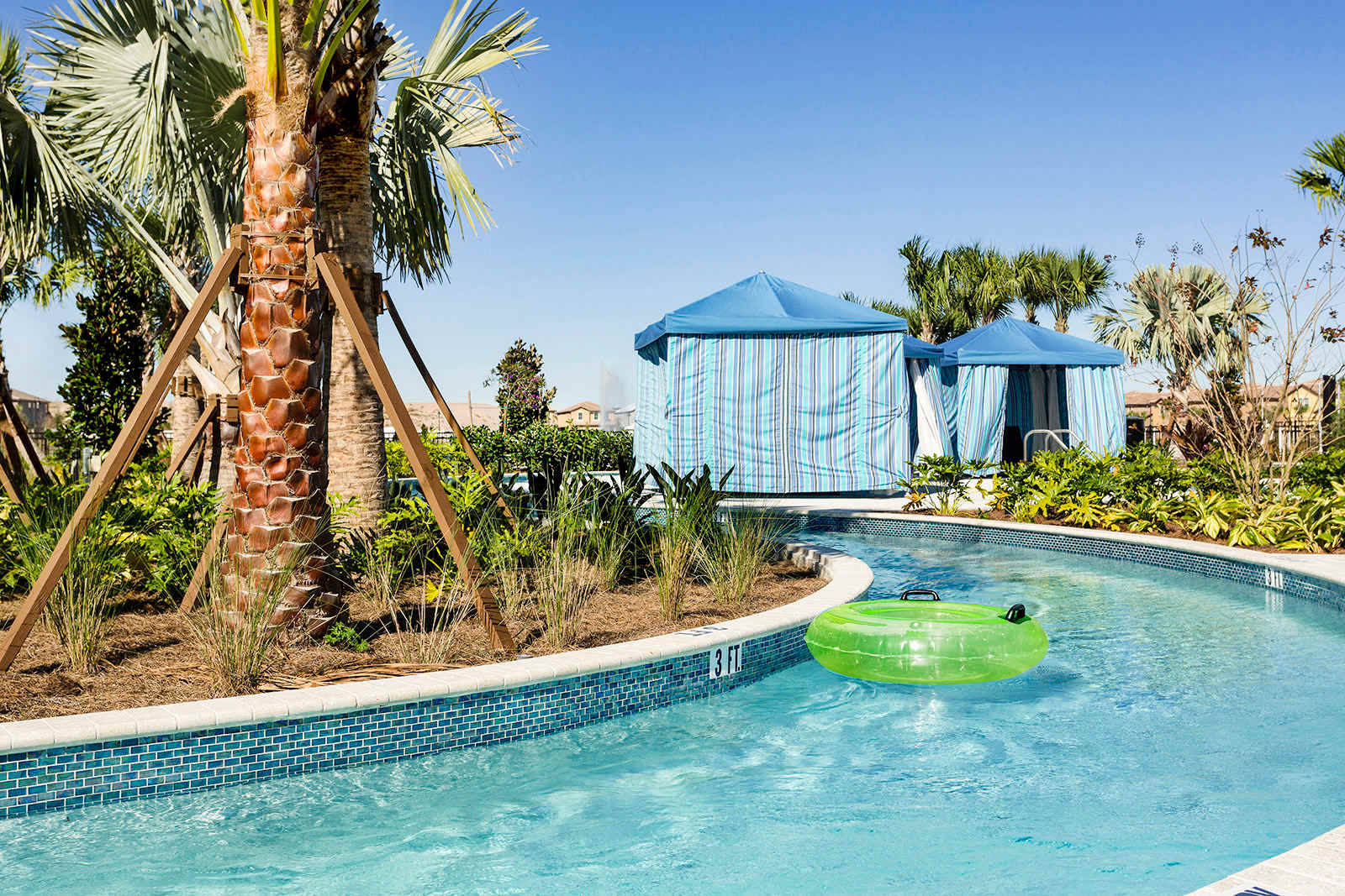 A winding lazy river at the resort pool area, featuring clear blue water and a green inflatable tube, surrounded by tropical palm trees and private cabanas under a bright, sunny sky.