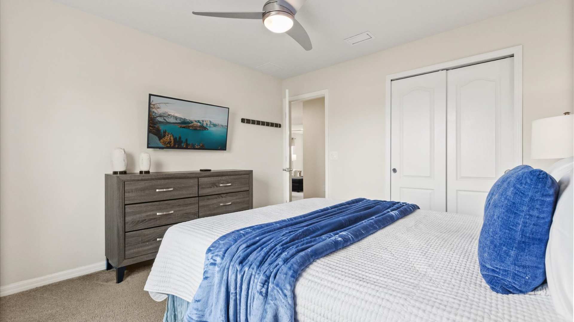 View of bedroom-3 showing a grey wooden dresser with a wall-mounted TV above it, and a closet with white bi-fold doors, complementing the blue and white bedding of the queen bed.