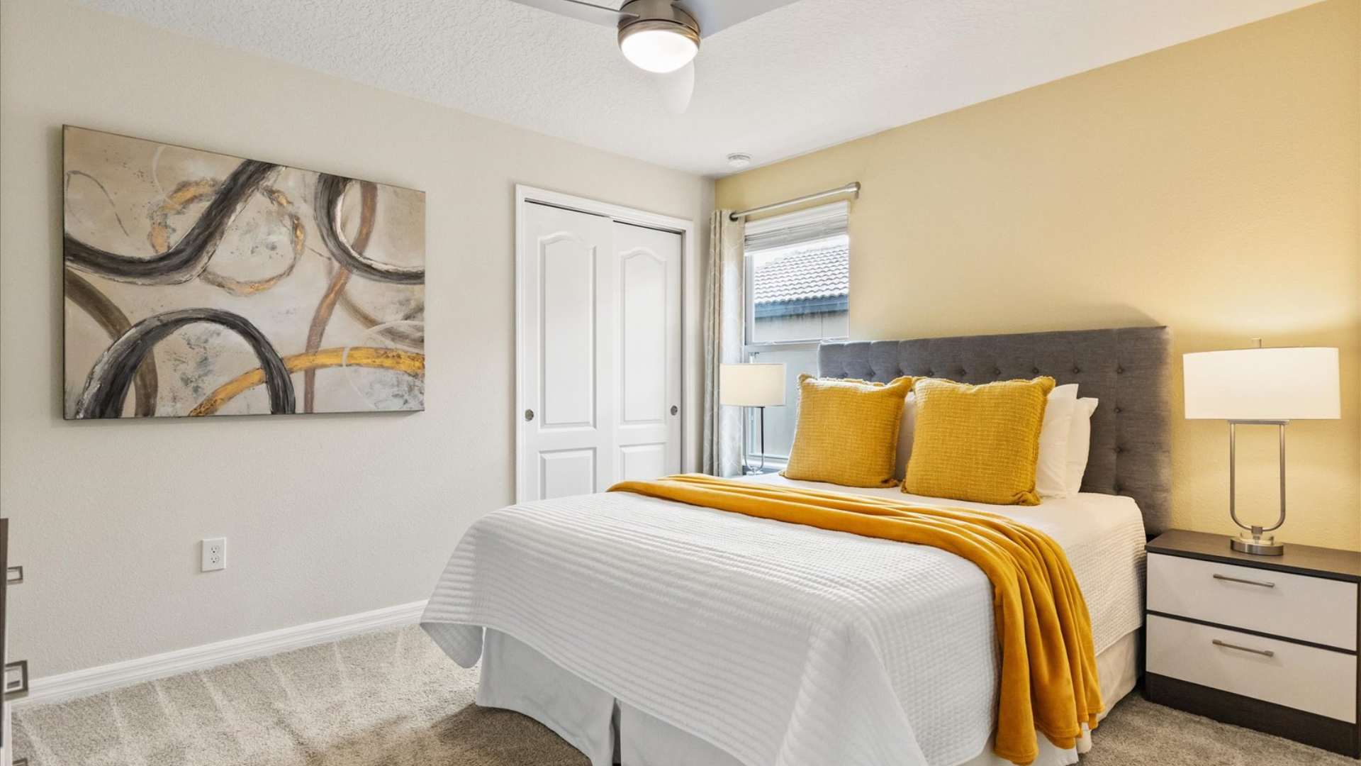 Cheerful bedroom with a queen-sized bed featuring a gray upholstered headboard and yellow accent pillows and throw, a dark nightstand, and a large abstract artwork on the wall.