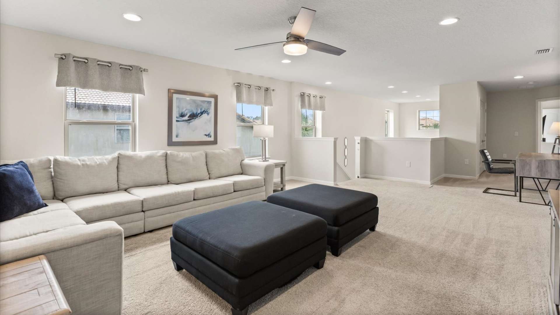 Spacious upstairs media room with light gray carpet, a large sectional sofa, two black ottomans, and an open railing overlooking the floor below, with multiple windows providing natural light.