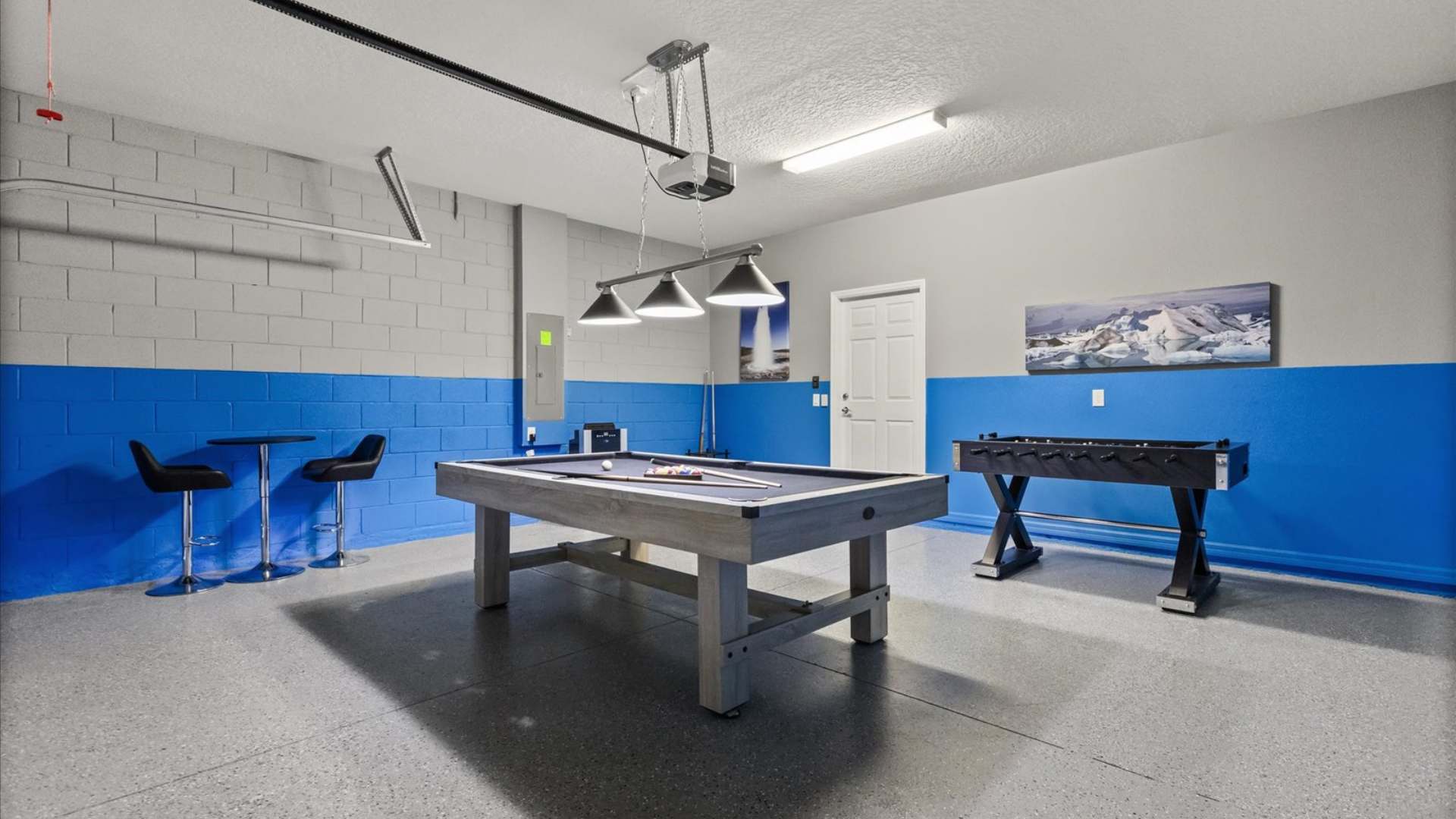 Another perspective of the game room, highlighting the pool table, foosball table, and a small high-top table with stools. The blue and gray painted walls and epoxy floor are prominent.
