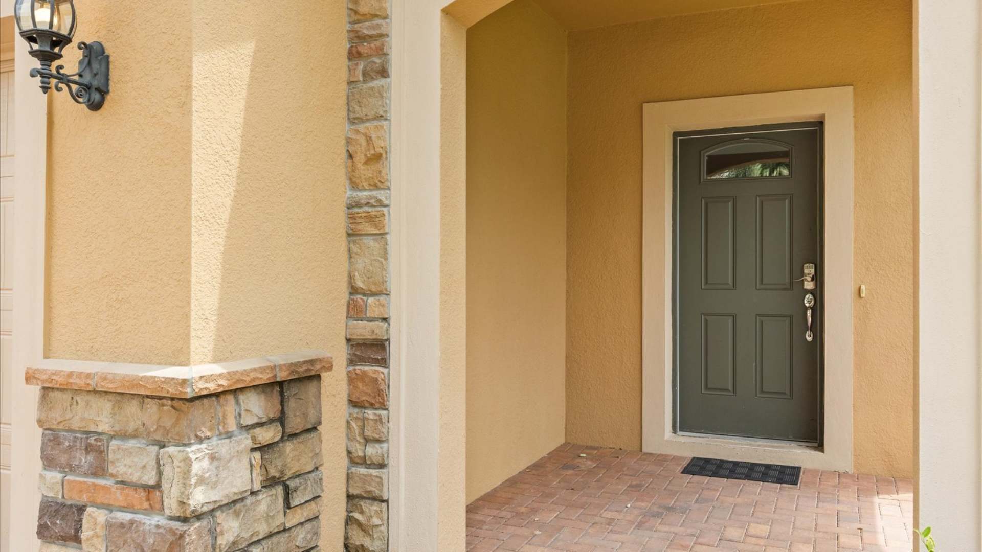 A welcoming front entrance featuring a dark green paneled door with a small window, set within warm yellow stucco walls and decorative stone accents, leading to a brick-patterned walkway.