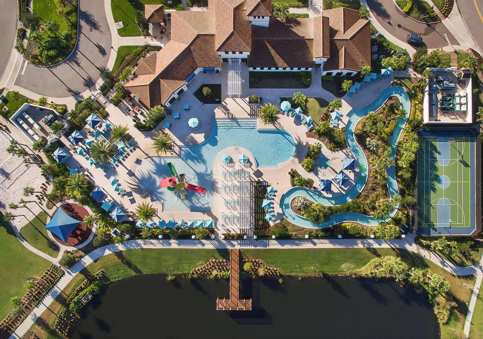 An expansive aerial view of a resort community, featuring a large swimming pool complex with a lazy river, water play area, sports court, and a lake with a pier, all surrounded by lush landscaping and residential buildings.