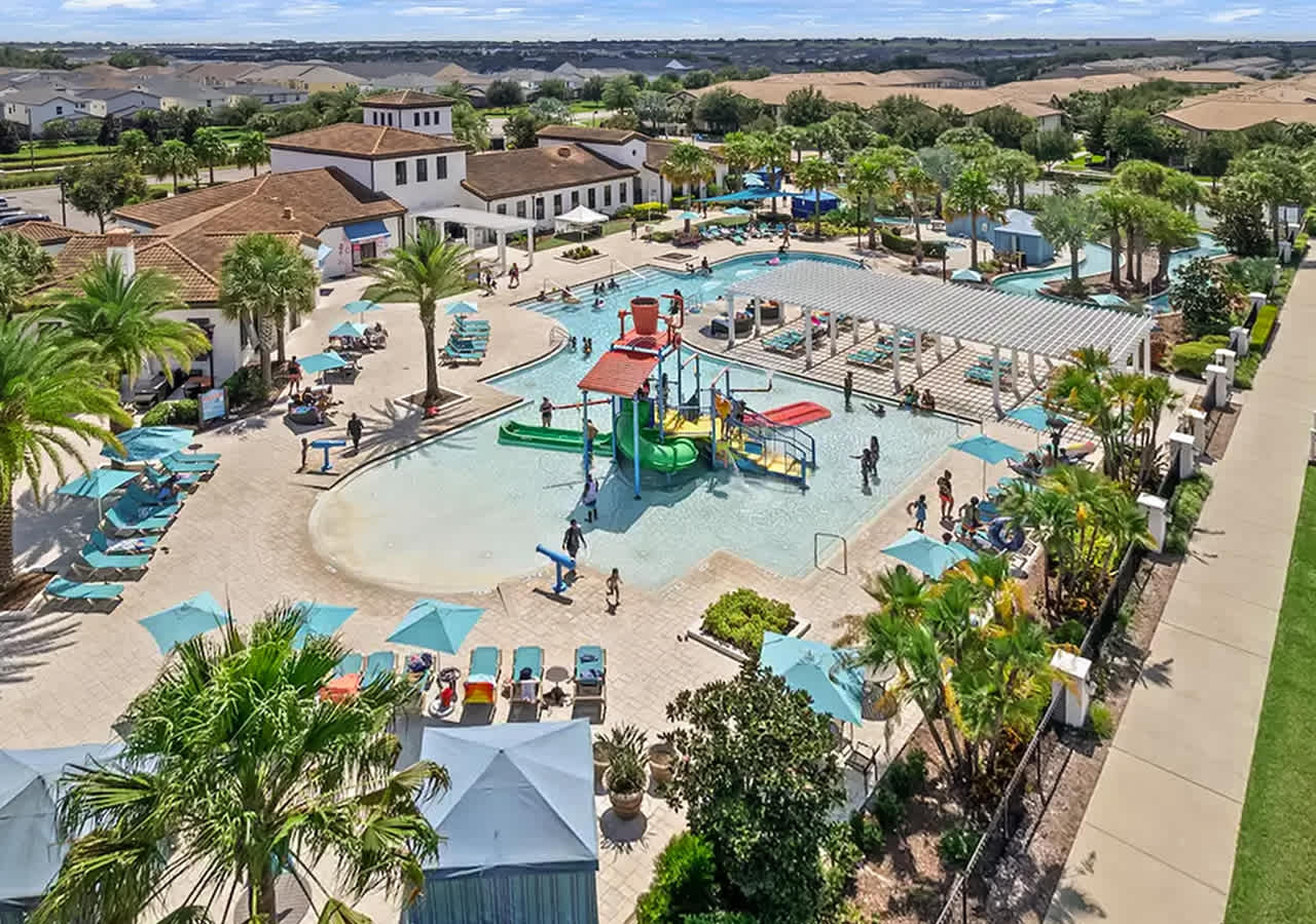 Aerial view of a sprawling resort amenity center, showcasing a large swimming pool complex with a water play area, a clubhouse, numerous lounge chairs, and surrounding residential properties under a clear sky.