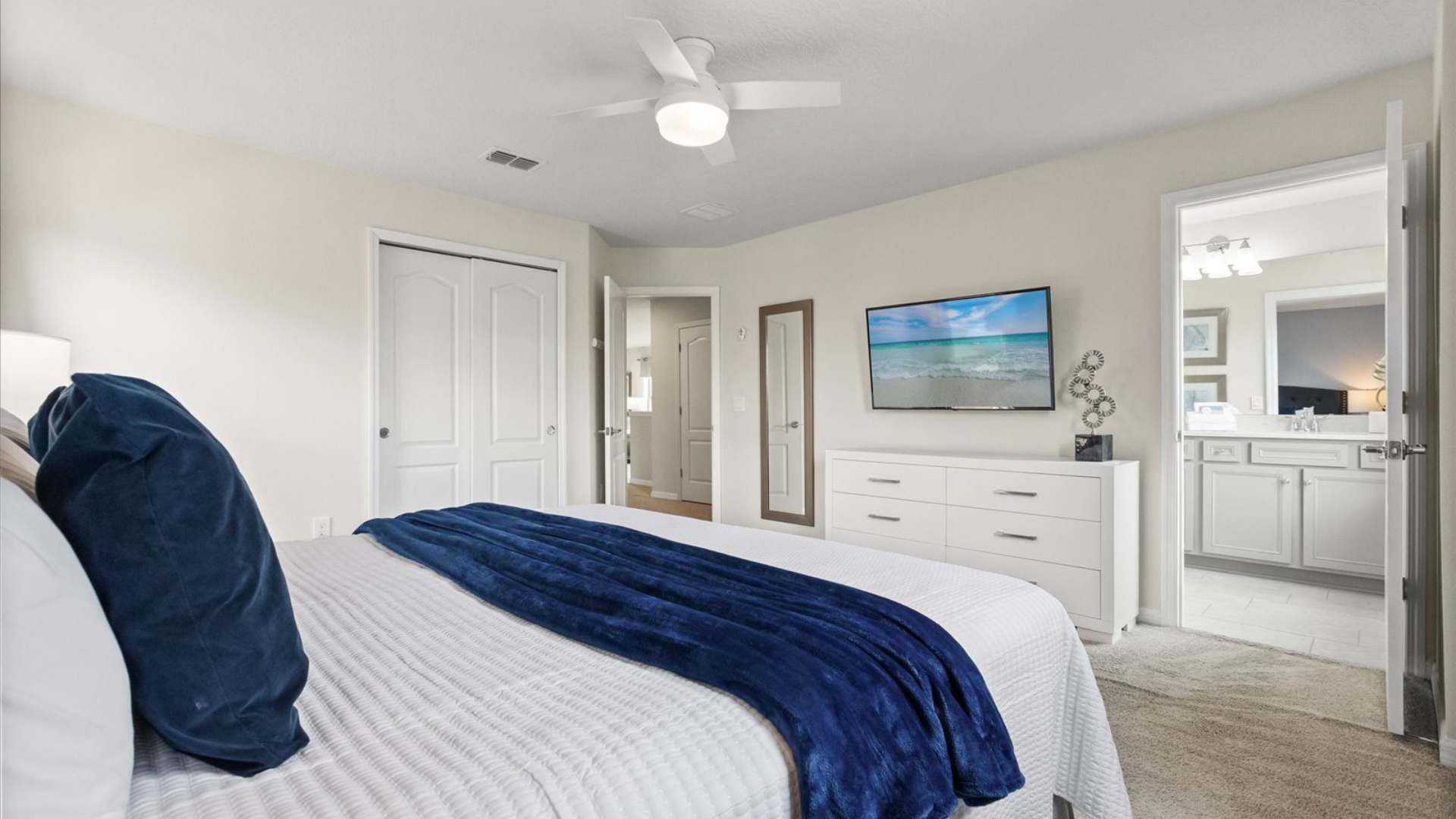 View of a bedroom showing a white dresser with a wall-mounted TV, a full-length mirror, and a bed with blue accents, with an open door leading to a bathroom.