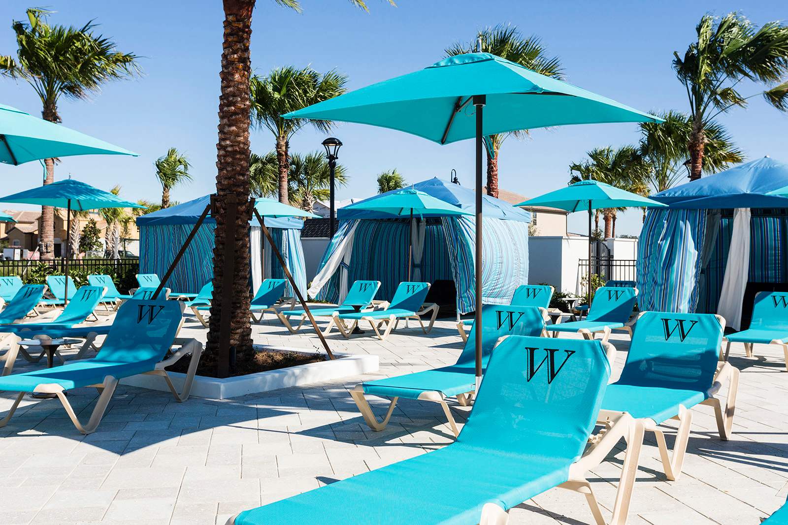 A vibrant poolside lounge area at the resort, featuring numerous turquoise lounge chairs with matching umbrellas and striped cabanas, set against a backdrop of palm trees and clear blue skies.