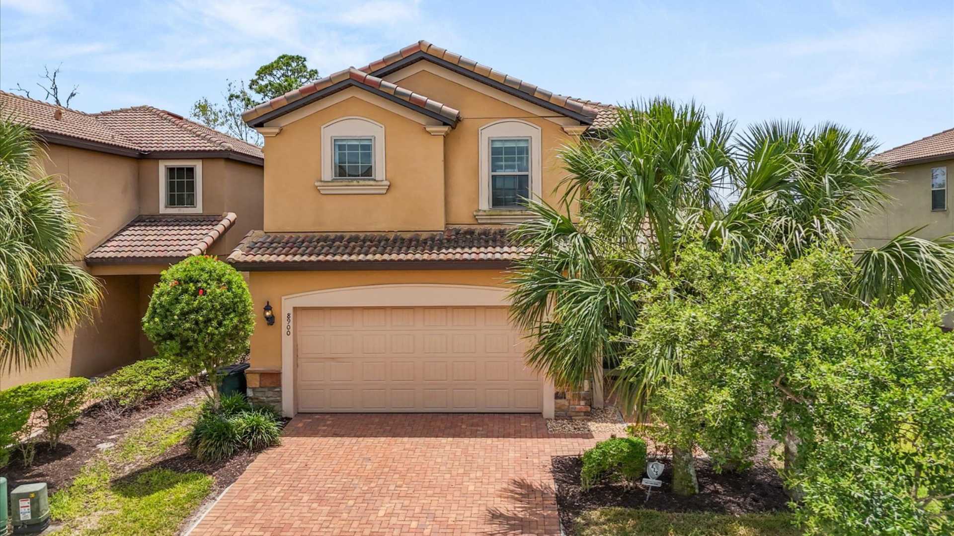 Front exterior of a two-story house with a warm yellow stucco facade, a red tile roof, and a two-car garage. Lush green landscaping with palm trees and shrubs frames the brick paver driveway.