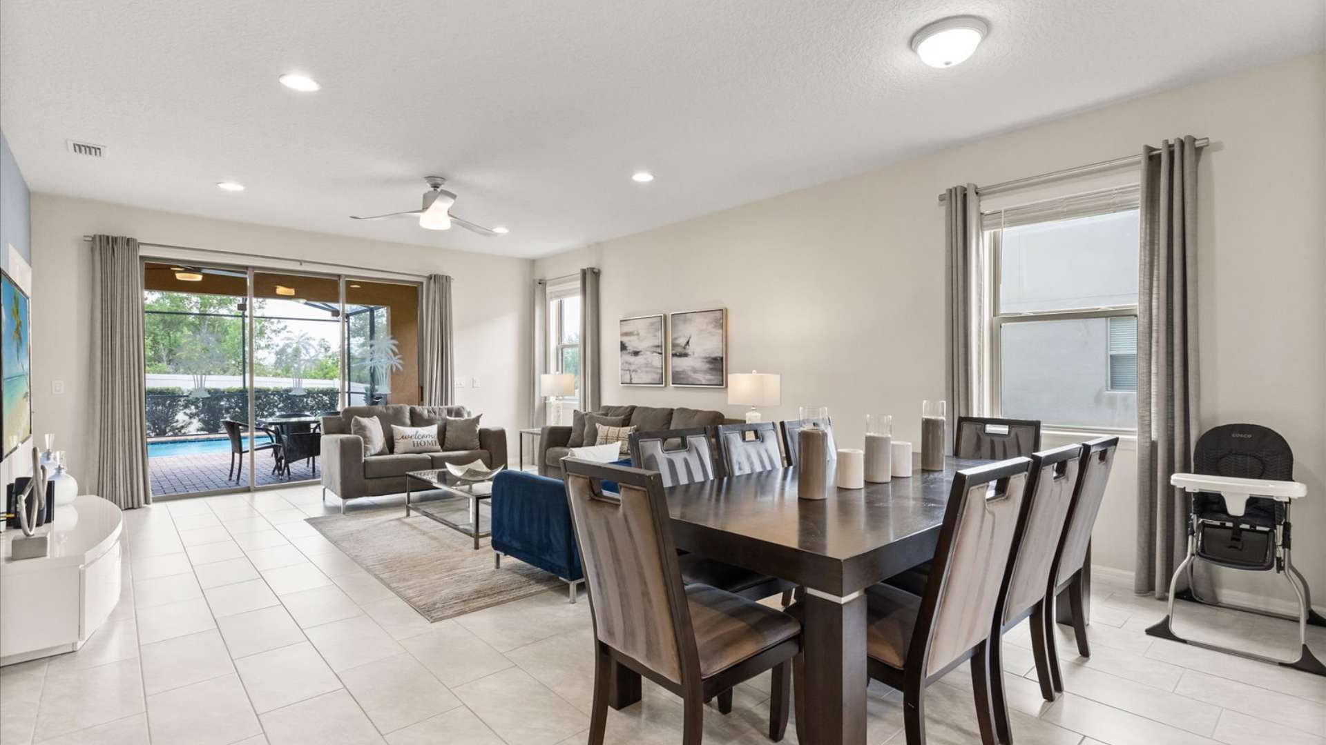 Open-concept living and dining area with light tile flooring, featuring gray sofas, a dark wood dining table with upholstered chairs, and sliding doors leading to a screened pool patio.