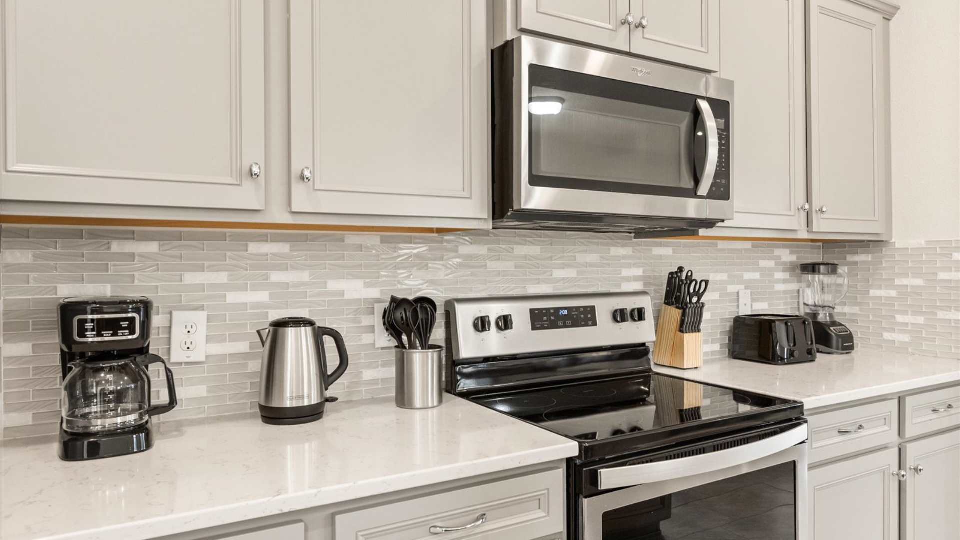 Detailed view of the kitchen counter with white quartz, light gray cabinets, a subway tile backsplash, and stainless steel appliances including a microwave, stove, coffee maker, and kettle.