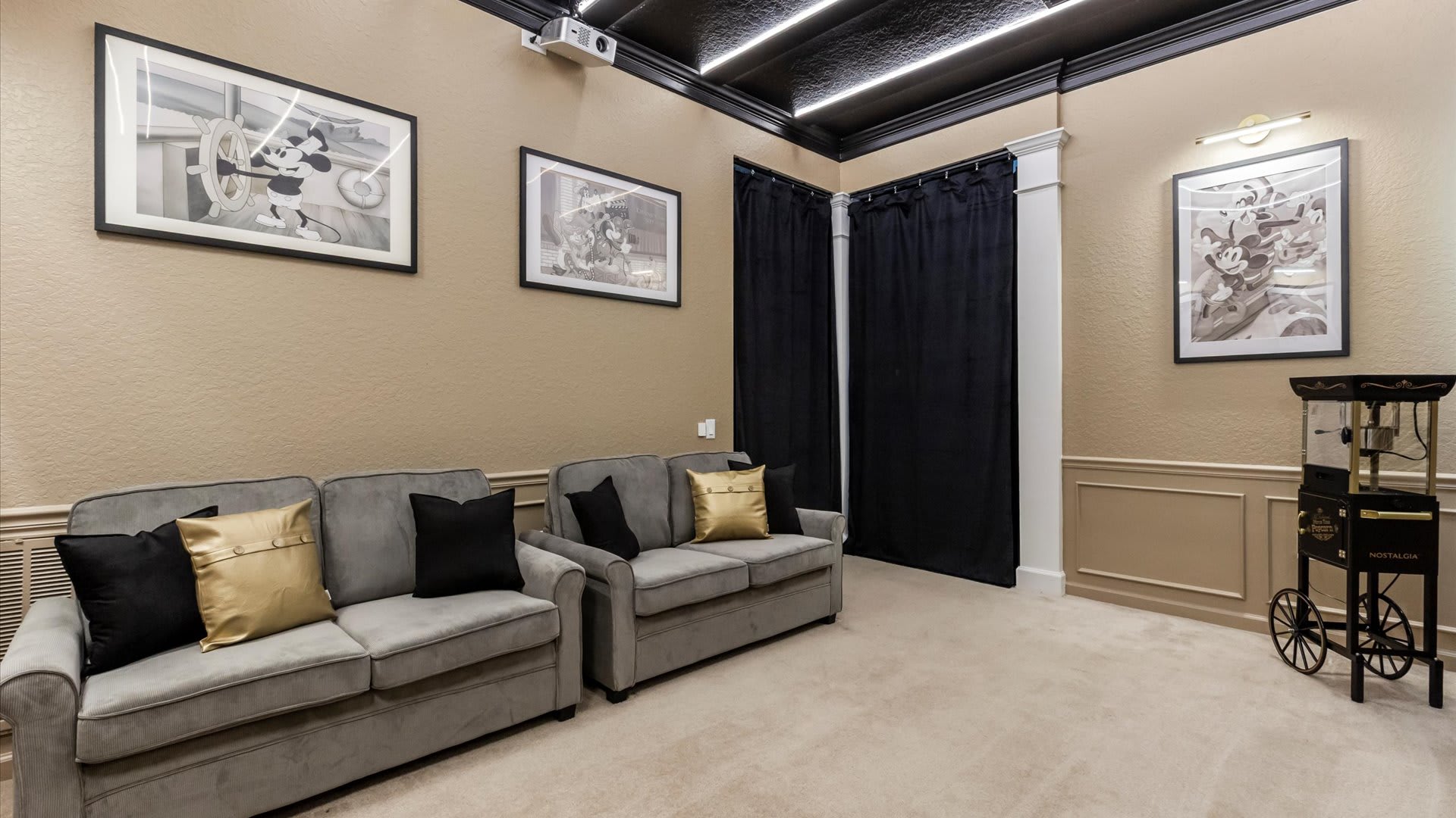 A wide shot of the media room, showcasing two grey sofas with decorative pillows, framed Mickey Mouse artwork, a ceiling-mounted projector, and black curtains in the corner, all within a themed space.