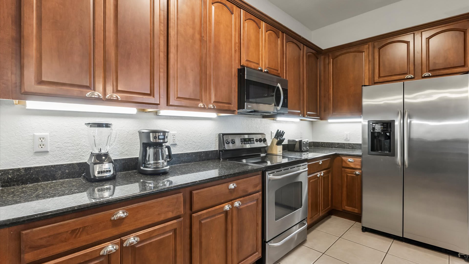 A close-up of the kitchen counter reveals a blender and coffee maker on black granite countertops, with warm wooden cabinets above and below. Under-cabinet lighting illuminates the appliances, and a stainless steel oven is visible in the background.