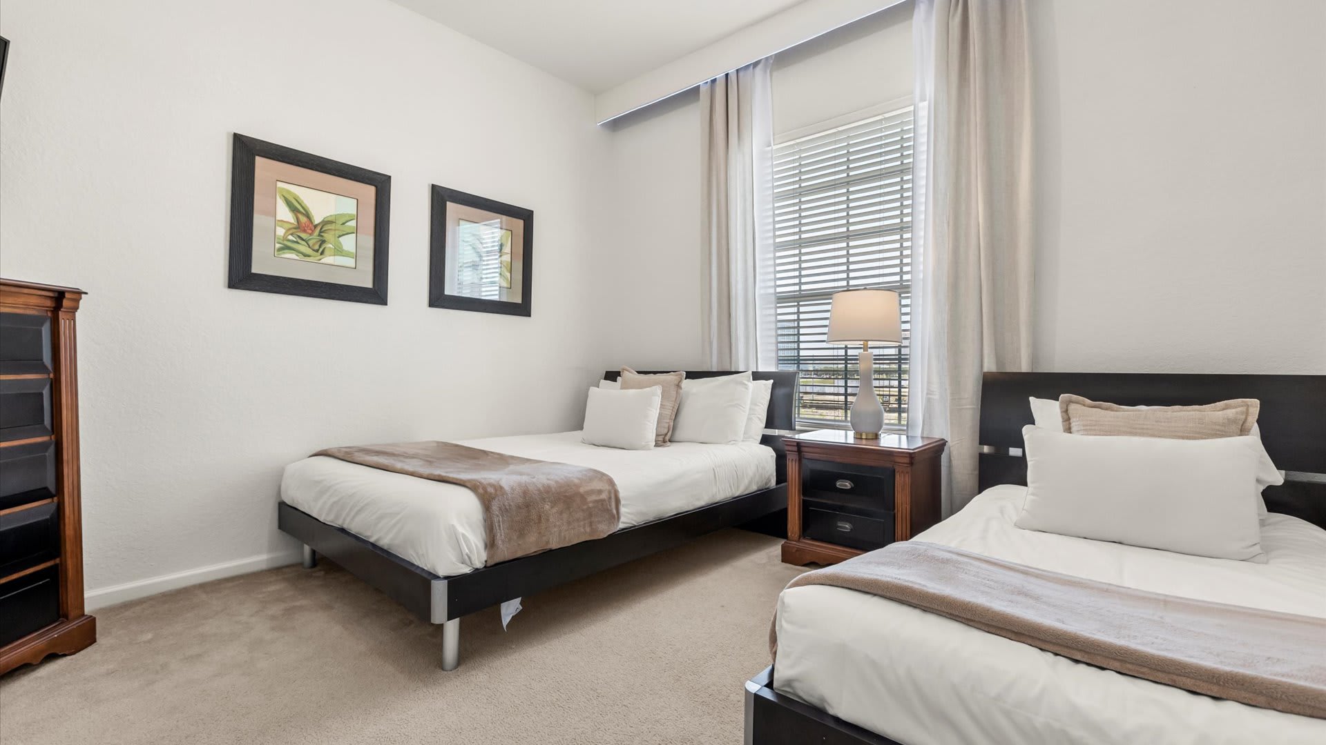 A bright bedroom furnished with two twin beds, each with white linens and a beige throw, separated by a dark wooden nightstand with a lamp. Two framed botanical prints hang on the wall, and a window with blinds and sheer curtains provides natural light.
