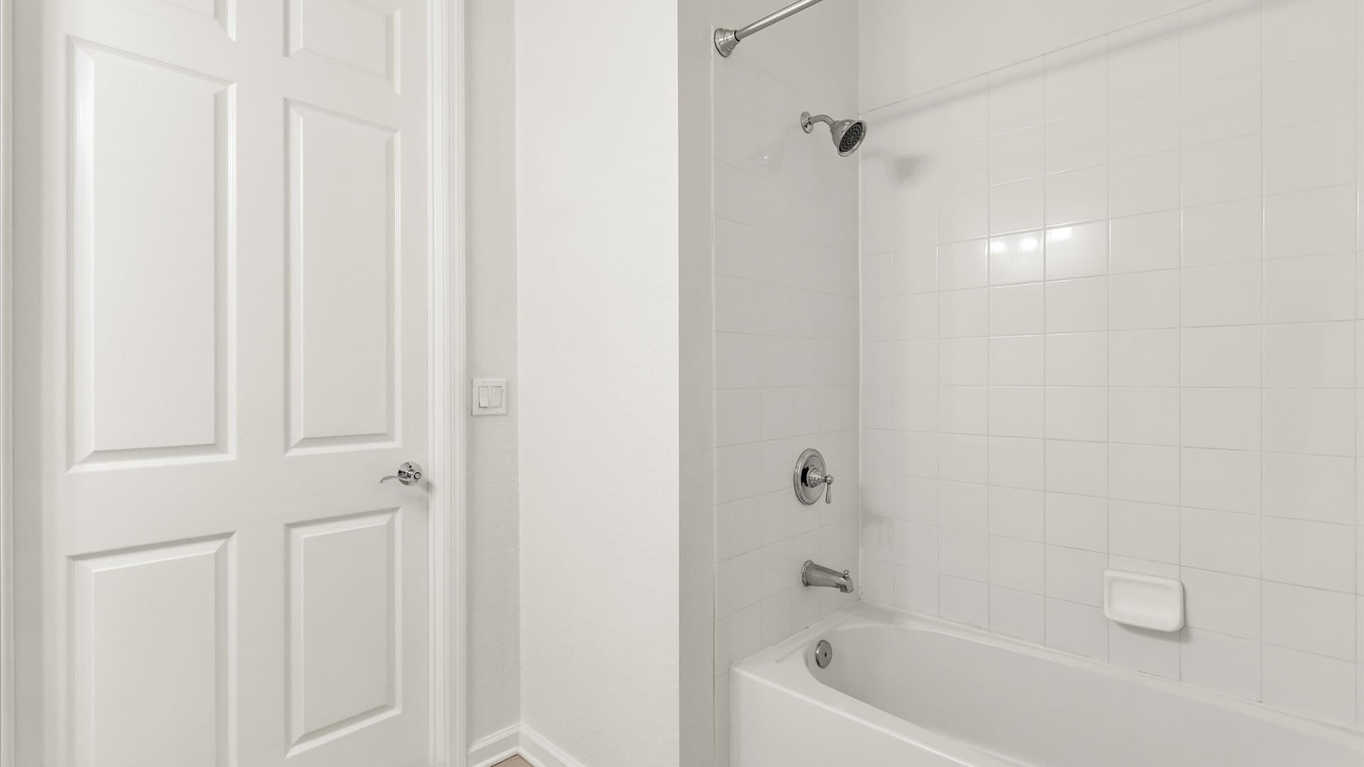 A clean white bathroom showcasing a shower-over-tub combination with white subway tiles and a chrome showerhead and fixtures. A white paneled door is visible to the left, suggesting an adjacent closet or another room.