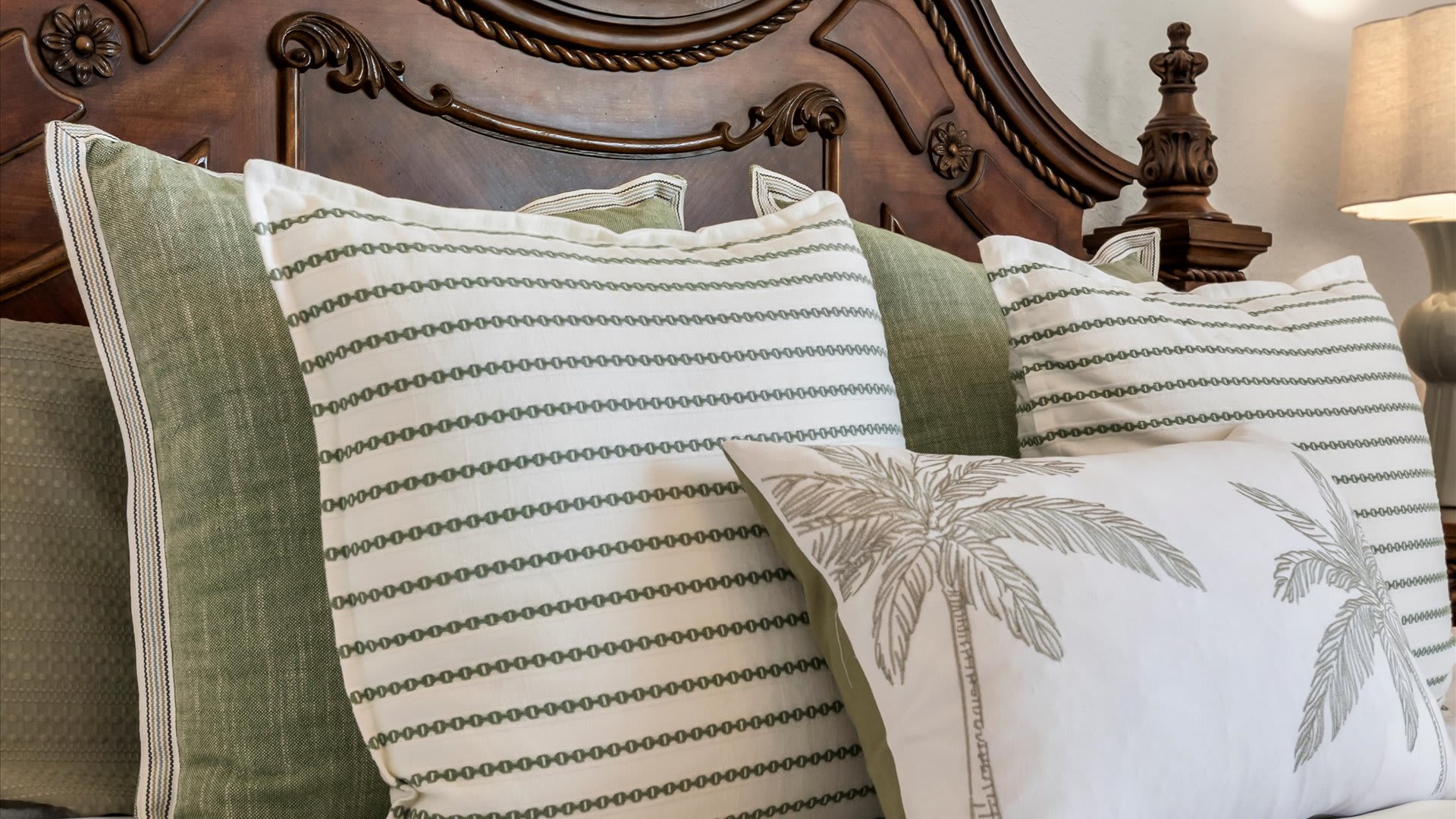 Close-up view of the bed in bedroom-1, showcasing decorative pillows in shades of green and white, including striped patterns and a palm tree motif, against the ornate dark wood headboard.