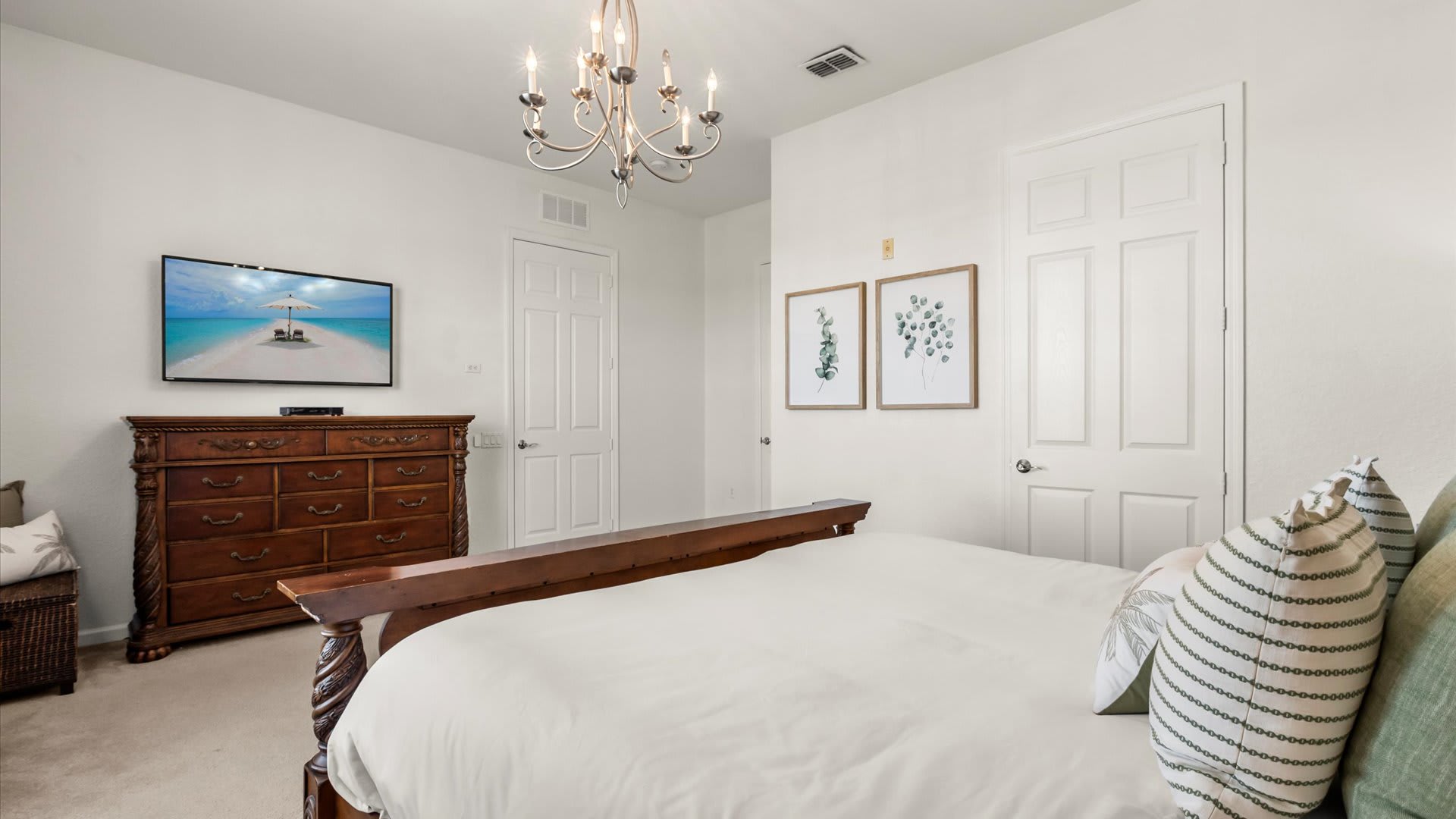 View of bedroom-1 from the foot of the bed, showing a large dark wood dresser with a flat-screen TV mounted above it, and two white doors. The room maintains its light carpet and white walls.