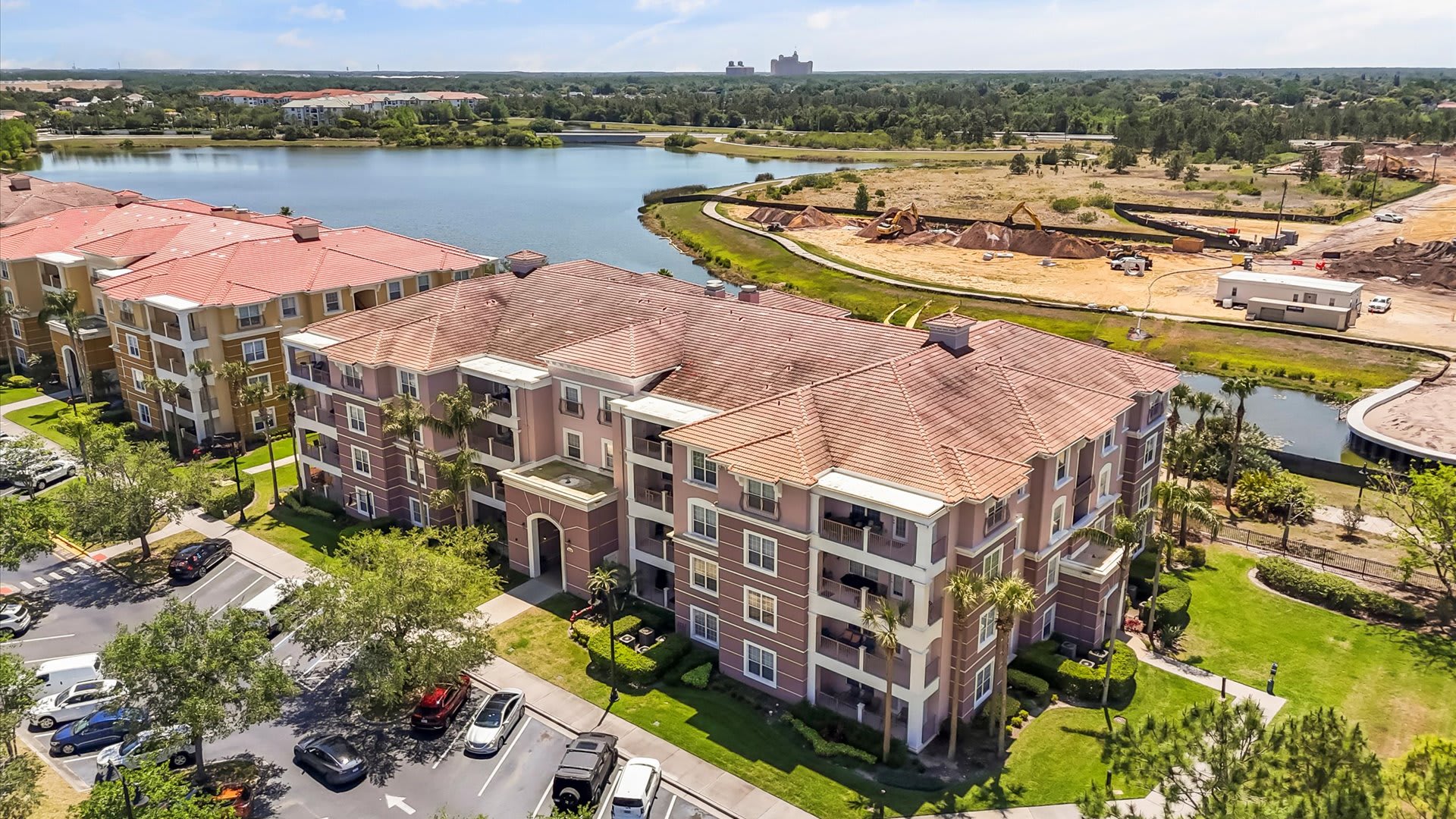 An aerial view of a large, multi-story condominium building with a terracotta-tiled roof, surrounded by lush green landscaping and a parking lot. A large lake and ongoing construction are visible in the background under a clear sky.