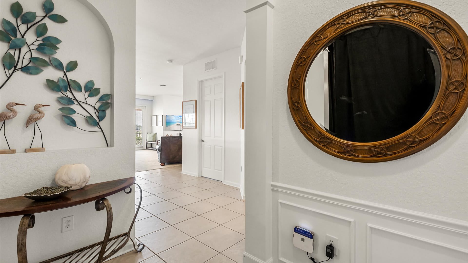 A bright foyer with light tile flooring, white walls, and a decorative console table with metal wall art, leading into a hallway with a glimpse of another room. A large round mirror with a woven frame hangs on the right wall.