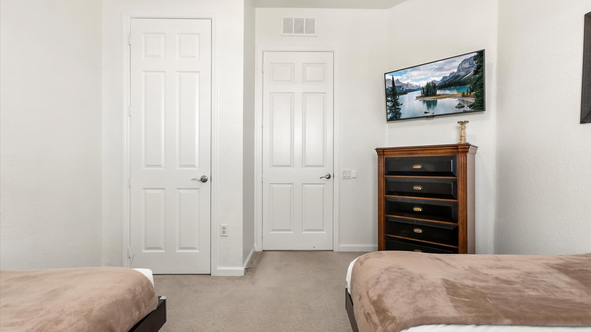 A view of the twin bedroom showing two white paneled doors, likely closets, and a wall-mounted TV above a dark wooden chest of drawers. The room has light-colored carpet and white walls.