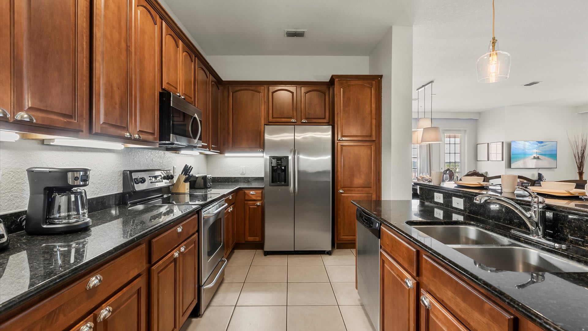 A full view of the kitchen showcases extensive warm wooden cabinetry, black granite countertops, and stainless steel appliances including a refrigerator, oven, microwave, and dishwasher. The floor is tiled, and under-cabinet lighting brightens the workspace.
