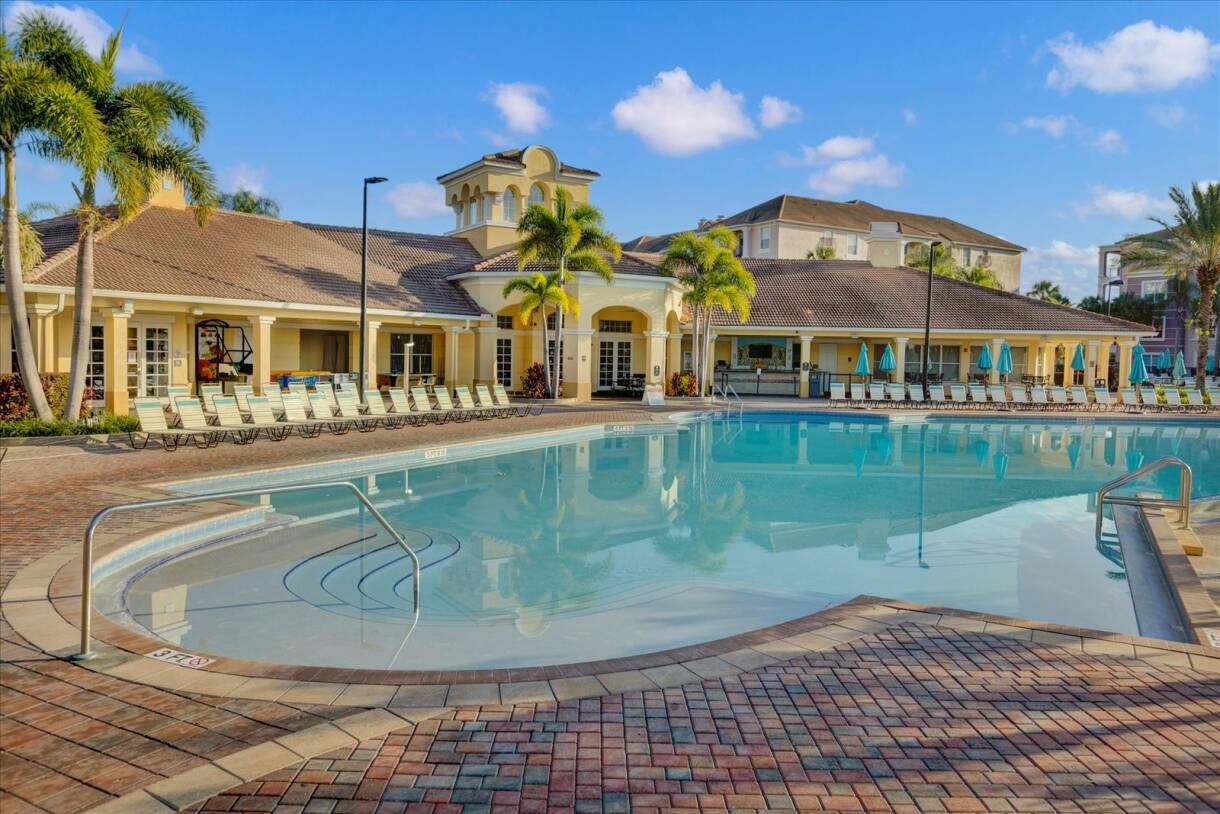 A large, inviting outdoor swimming pool surrounded by numerous lounge chairs and palm trees, with a grand clubhouse building visible in the background under a clear blue sky.