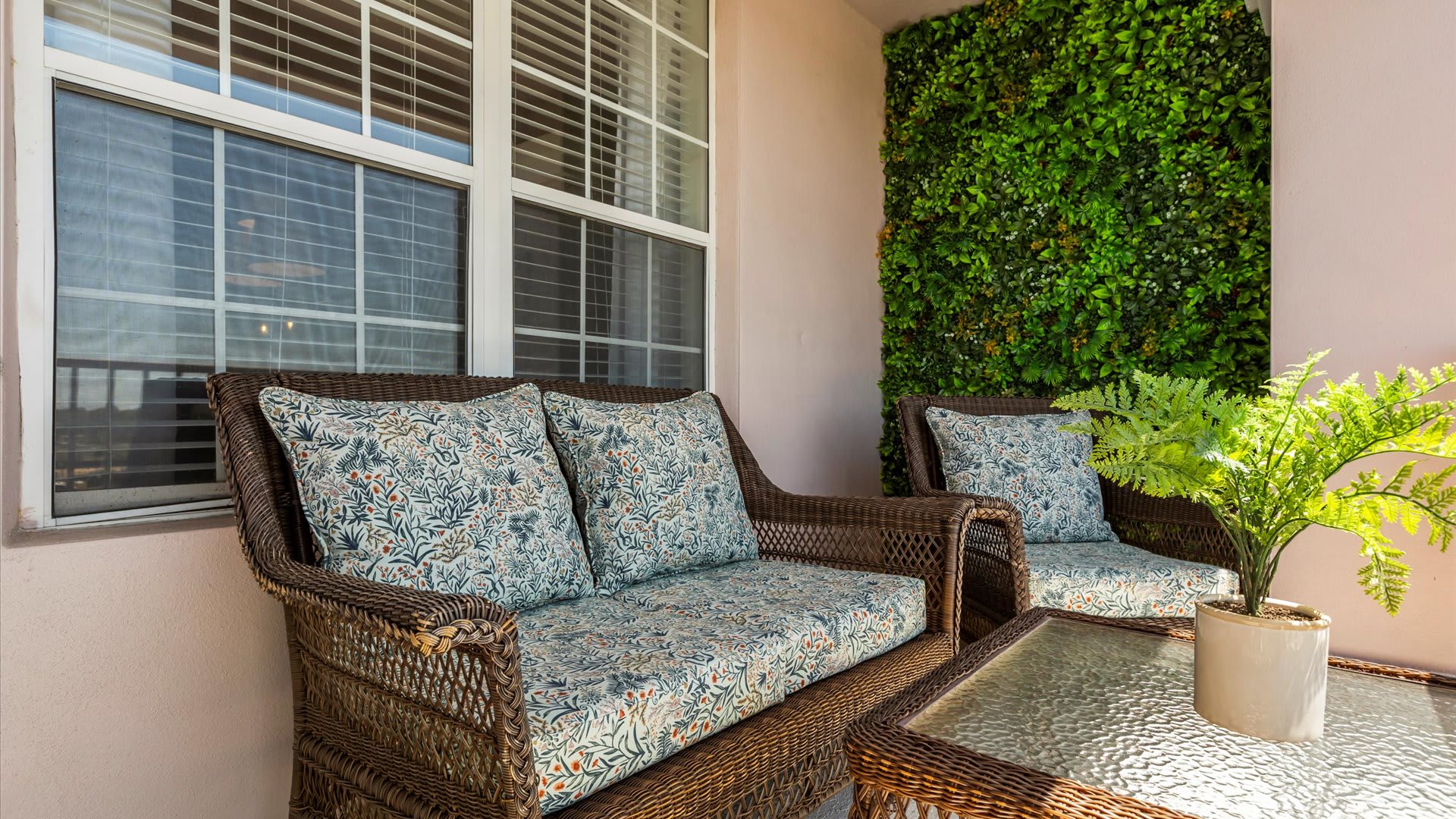 A cozy corner of a balcony featuring a wicker sofa and chair with patterned cushions, a small glass-top table, and a lush green wall, providing a comfortable outdoor seating area.