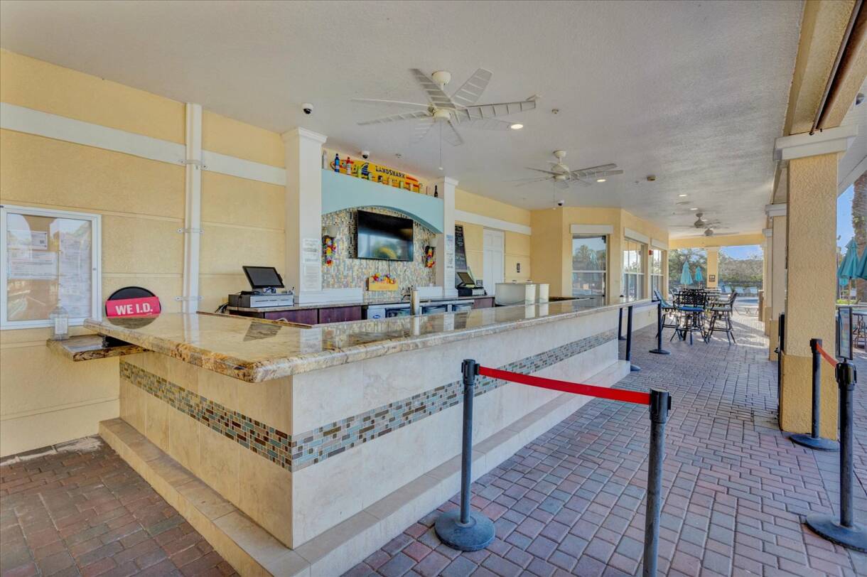 An outdoor bar with a long granite countertop and mosaic tile facade, featuring ceiling fans, a mounted TV, and a "WE'LD" sign, set on a brick paver patio.