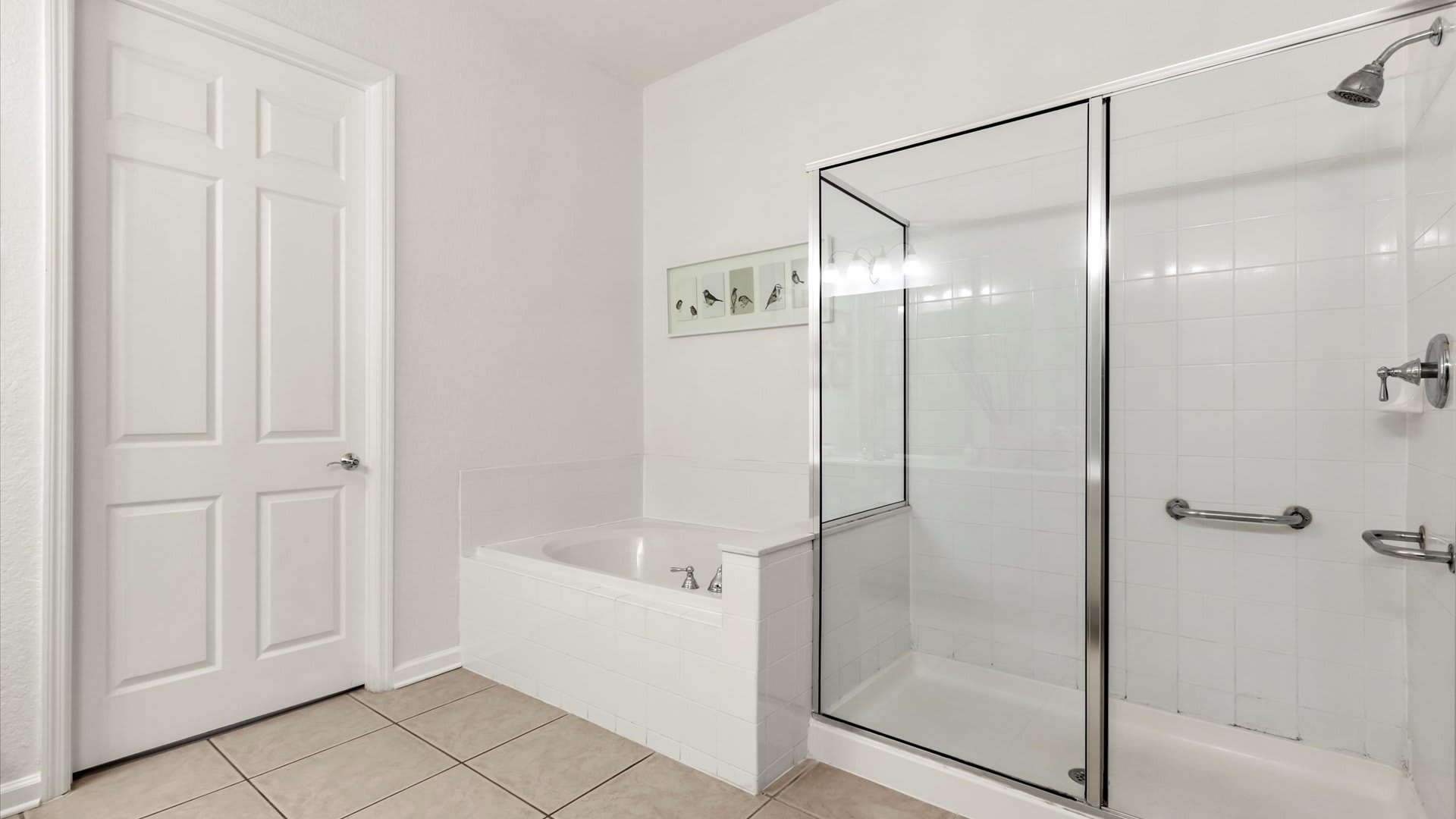 Close-up of the white built-in bathtub and the adjacent walk-in shower with a glass door and white tiled walls in bathroom-1, featuring light tile flooring and a white door.