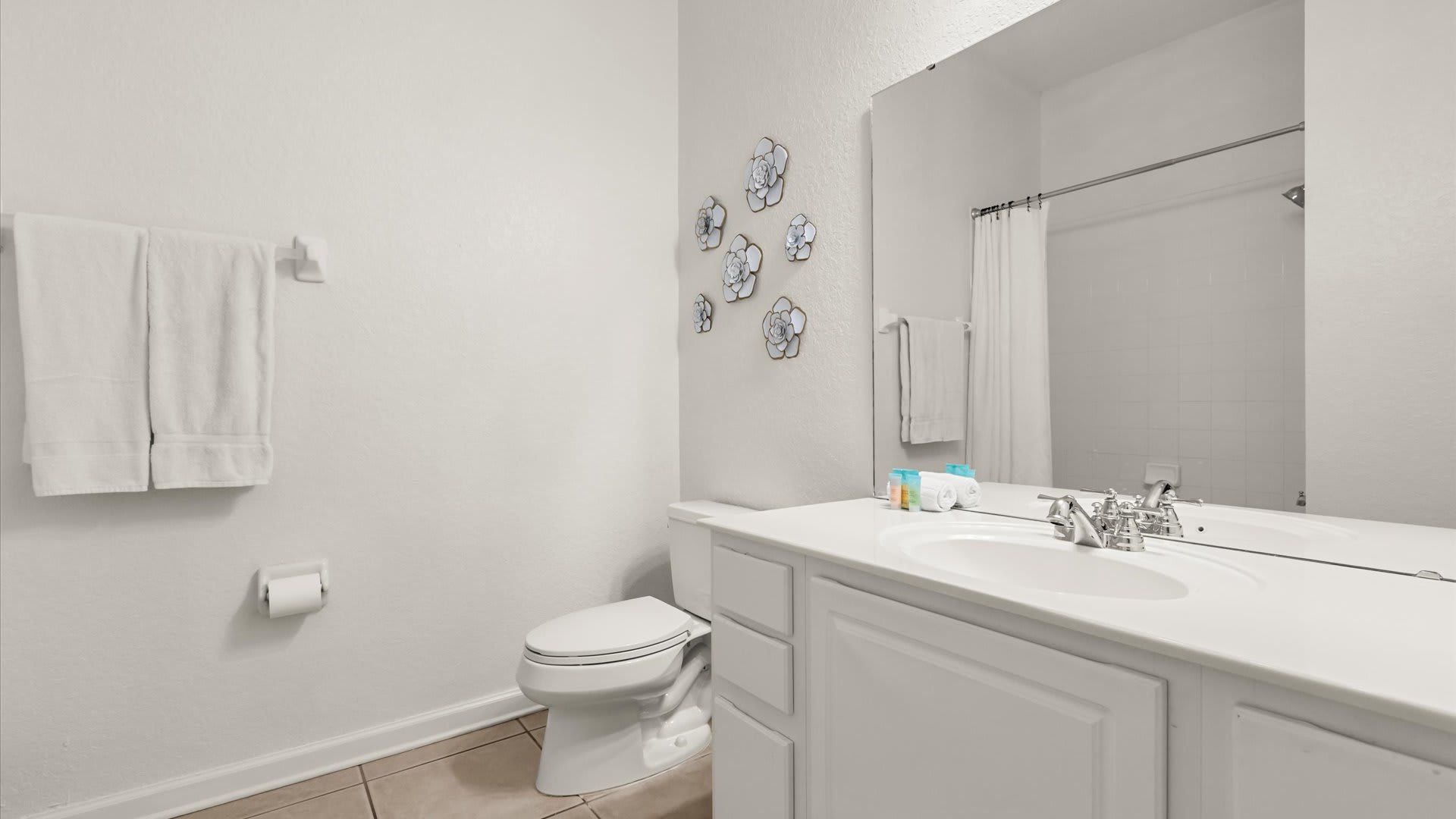 A bright bathroom featuring a white vanity with a single sink, a large mirror, and a toilet. White towels are neatly folded on a rack, and decorative floral wall art adorns the wall above the toilet.
