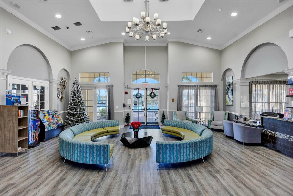 A spacious and bright lobby with high ceilings, a large modern chandelier, and two curved blue and green striped sofas arranged around a dark coffee table, with a Christmas tree in the corner.