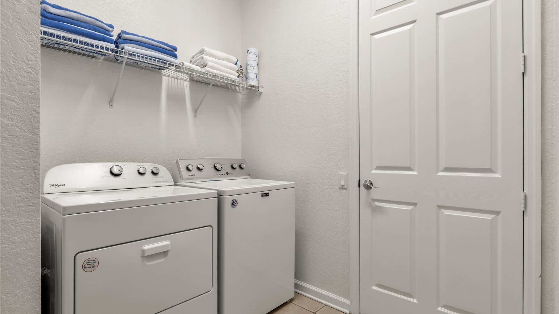 A functional laundry room equipped with a white Whirlpool washing machine and a Maytag dryer, positioned beneath a wire shelf stocked with neatly folded white and blue towels. A white paneled door is visible to the right.