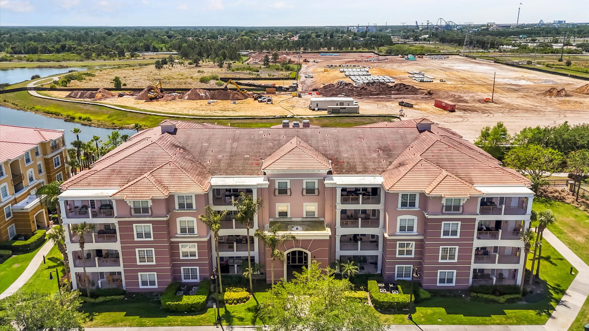 A high-angle aerial shot showcasing the full facade of a large, multi-story condominium complex with numerous balconies and a distinctive terracotta roof. The building overlooks a lake, with a large construction area visible in the distance.