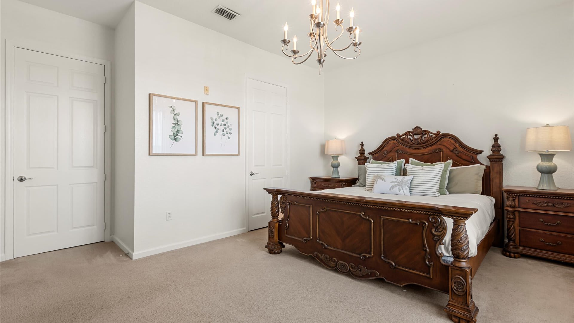 Spacious bedroom featuring a grand, ornate dark wood king-sized bed with a tall headboard, flanked by matching nightstands with lamps, under a classic chandelier. The room has light carpet and white walls with two framed botanical prints.