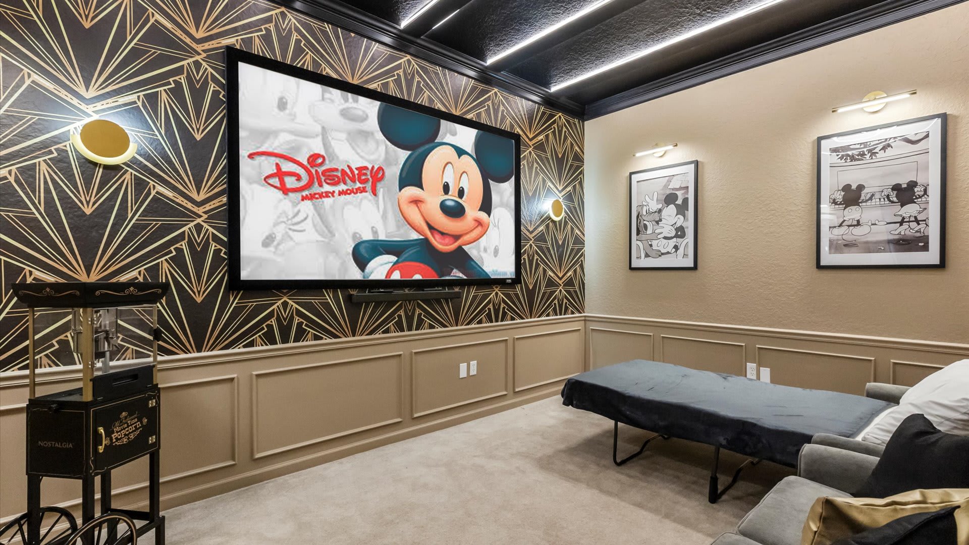 The media room showcases a large TV displaying Mickey Mouse, set against a striking black and gold geometric wallpaper. A popcorn machine stands to the left, complementing the grey sofa and pull-out bed.