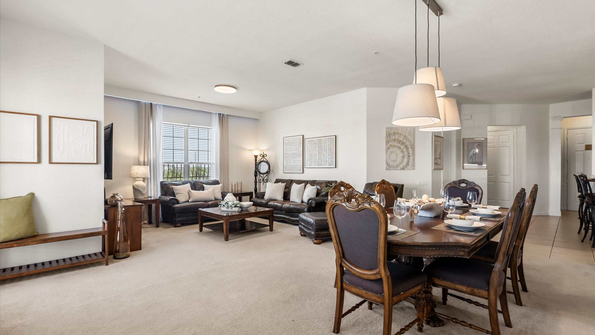 An open-concept living and dining area features brown leather sofas and a coffee table in the living space, transitioning to a dark wood dining table with ornate chairs. Pendant lights hang above the dining table, and light-colored carpet covers the floor.
