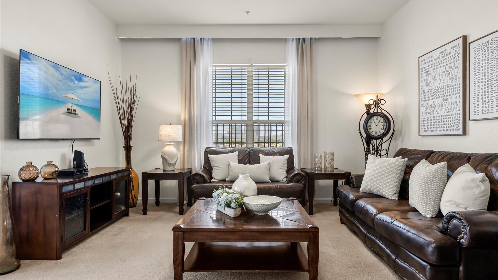 A spacious living room is furnished with two brown leather sofas adorned with white pillows, a dark wood coffee table, and a large wall-mounted TV. A unique floor lamp with a clock adds character, with light-colored blinds covering the windows.