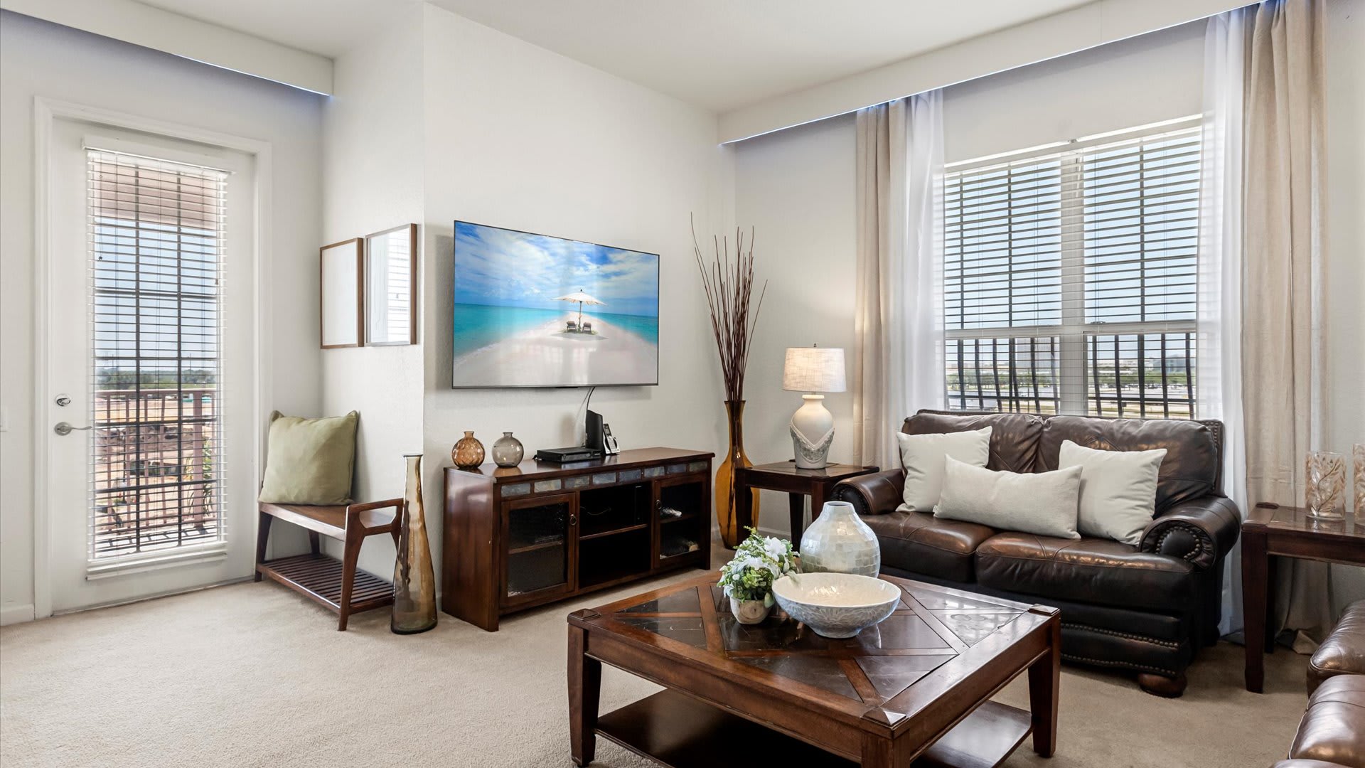 A bright living room features a large wall-mounted TV displaying a beach scene, a brown leather sofa with white pillows, and a dark wood coffee table. A glass door opens to an outdoor space, with light-colored blinds on the windows.
