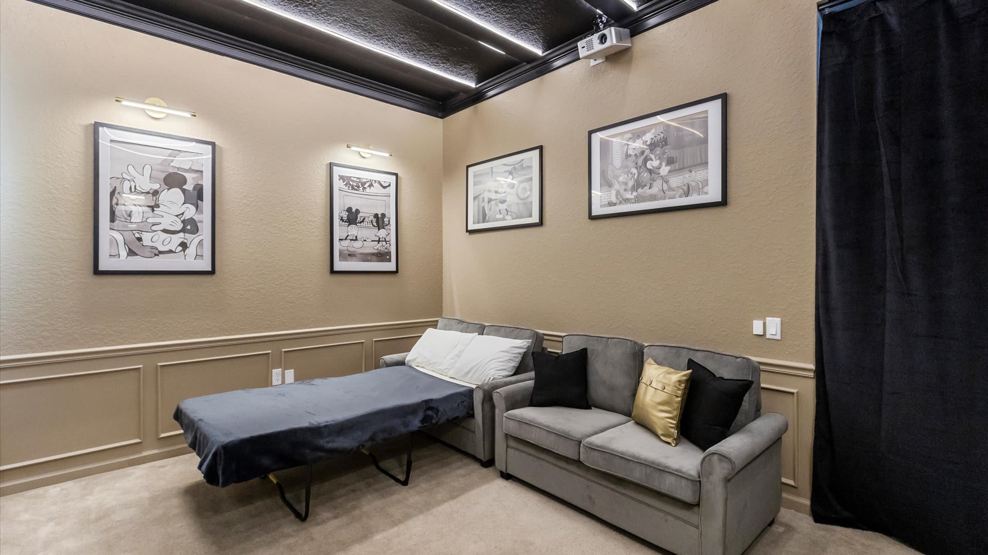 A cozy media room features a dark ceiling with linear lighting, beige walls with wainscoting, and framed black and white art. A grey sofa and a pull-out bed provide seating, with a projector visible on the ceiling.