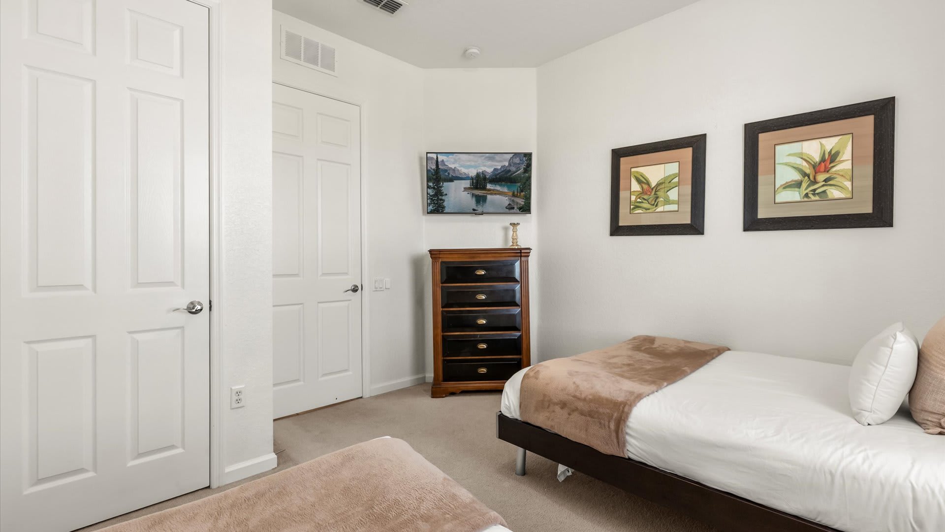 A corner view of the twin bedroom, highlighting the two beds with white bedding and beige throws, and a wall-mounted television. Two framed botanical artworks are visible on the wall, adding a touch of decor.