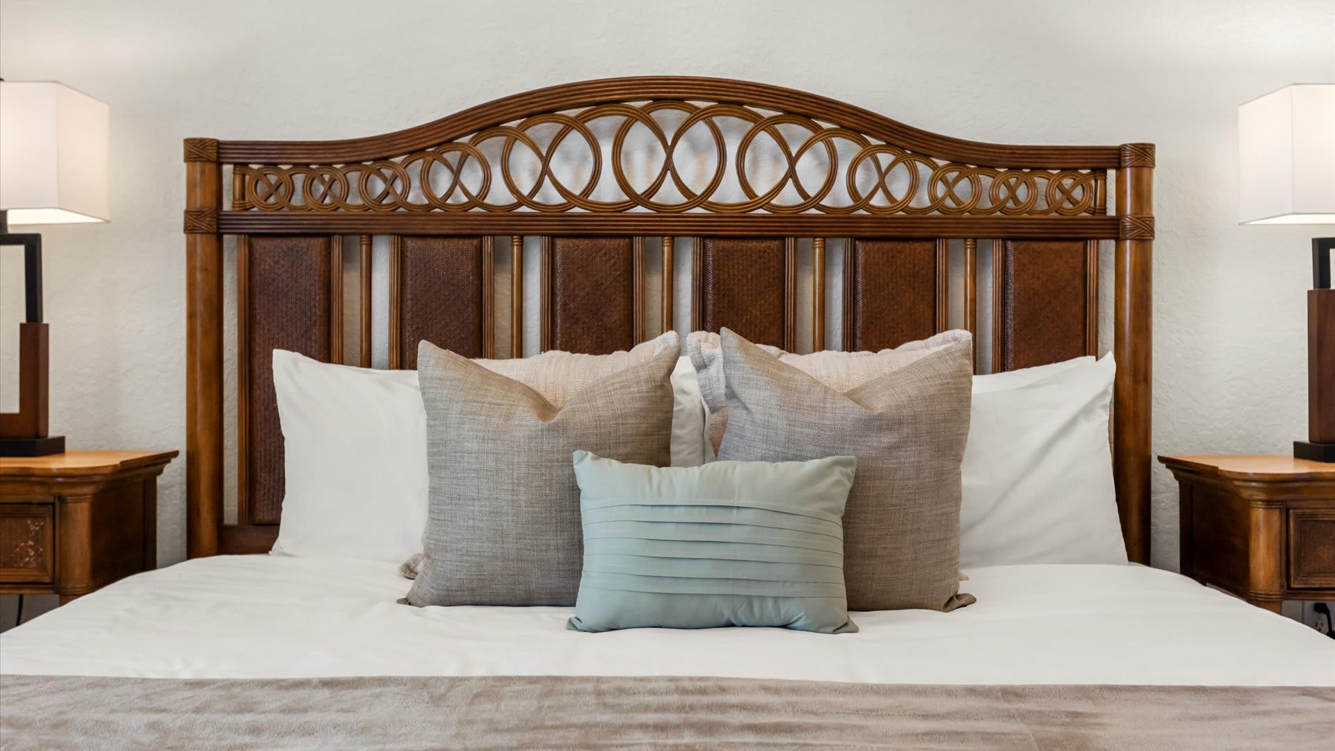 A close-up view of a queen or king-sized bed with a decorative wooden headboard featuring intricate circular patterns. The bed is made with white linens, a beige throw, and several pillows in shades of beige and light blue, flanked by two wooden nightstands with lamps.