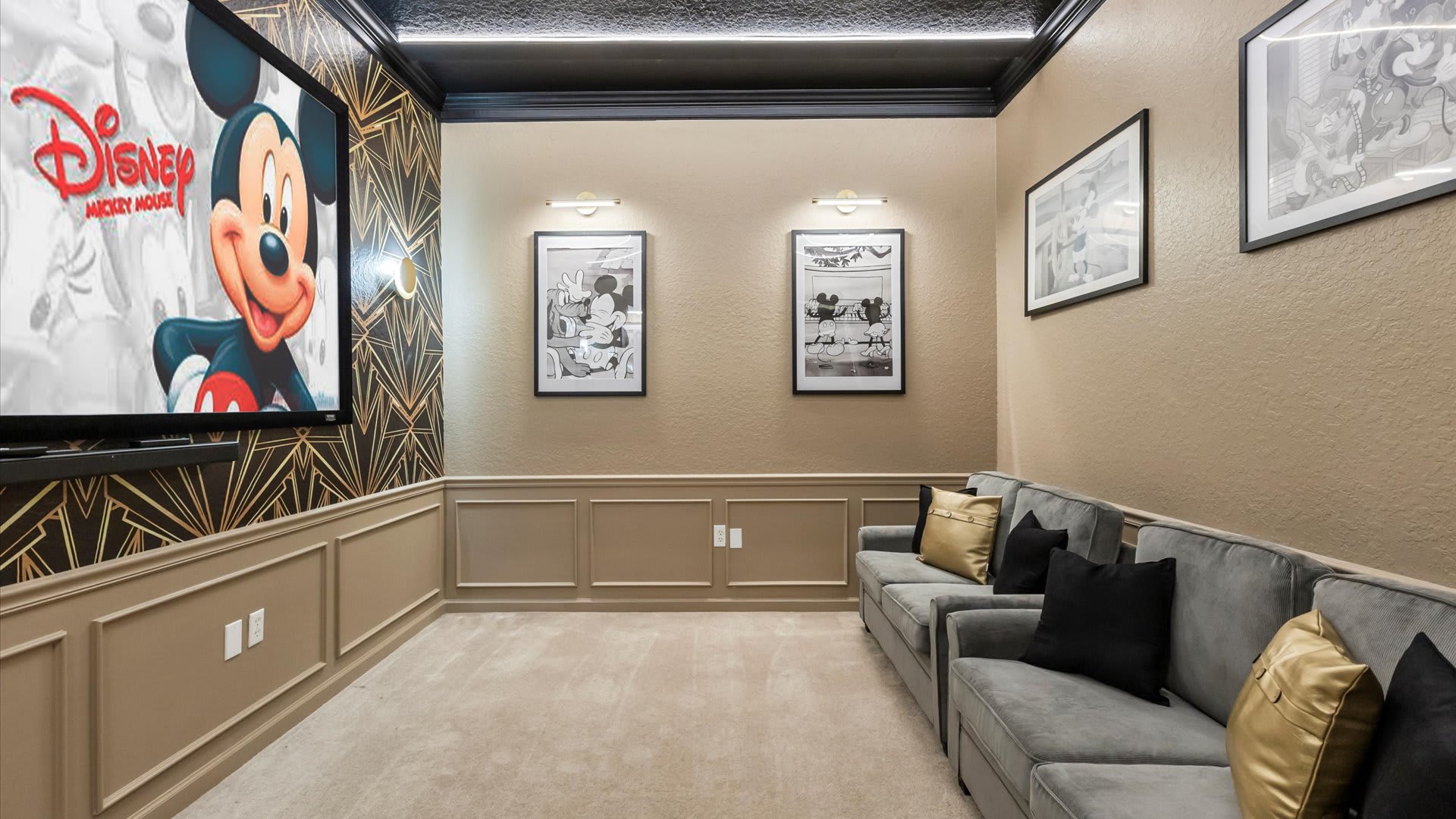 The media room's seating area, featuring two comfortable grey sofas adorned with black and gold throw pillows, set against a wall decorated with framed Mickey Mouse artwork and wainscoting.