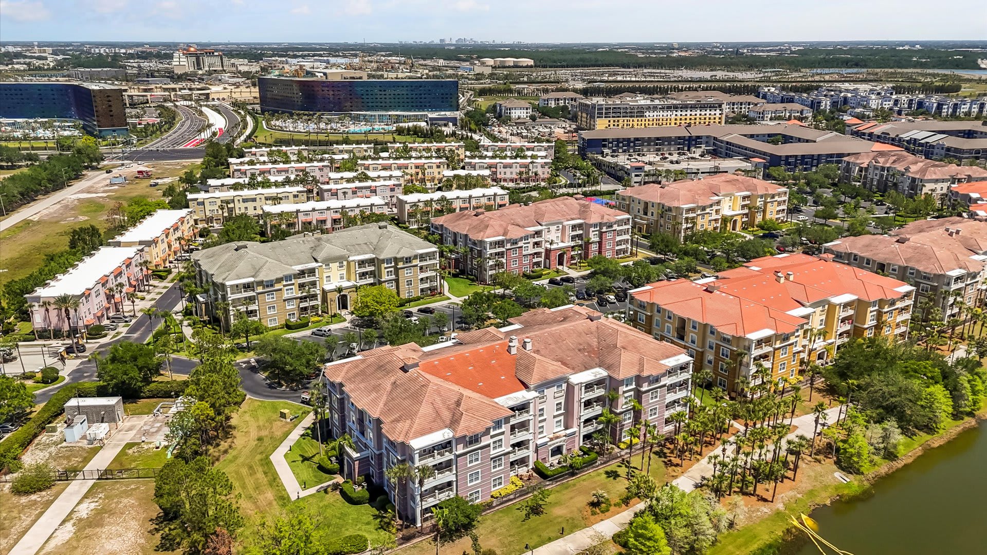 An aerial view of a large condominium complex with multiple multi-story buildings featuring various colored facades and red tile roofs, surrounded by green landscaping and a lake, with a city skyline in the distance.