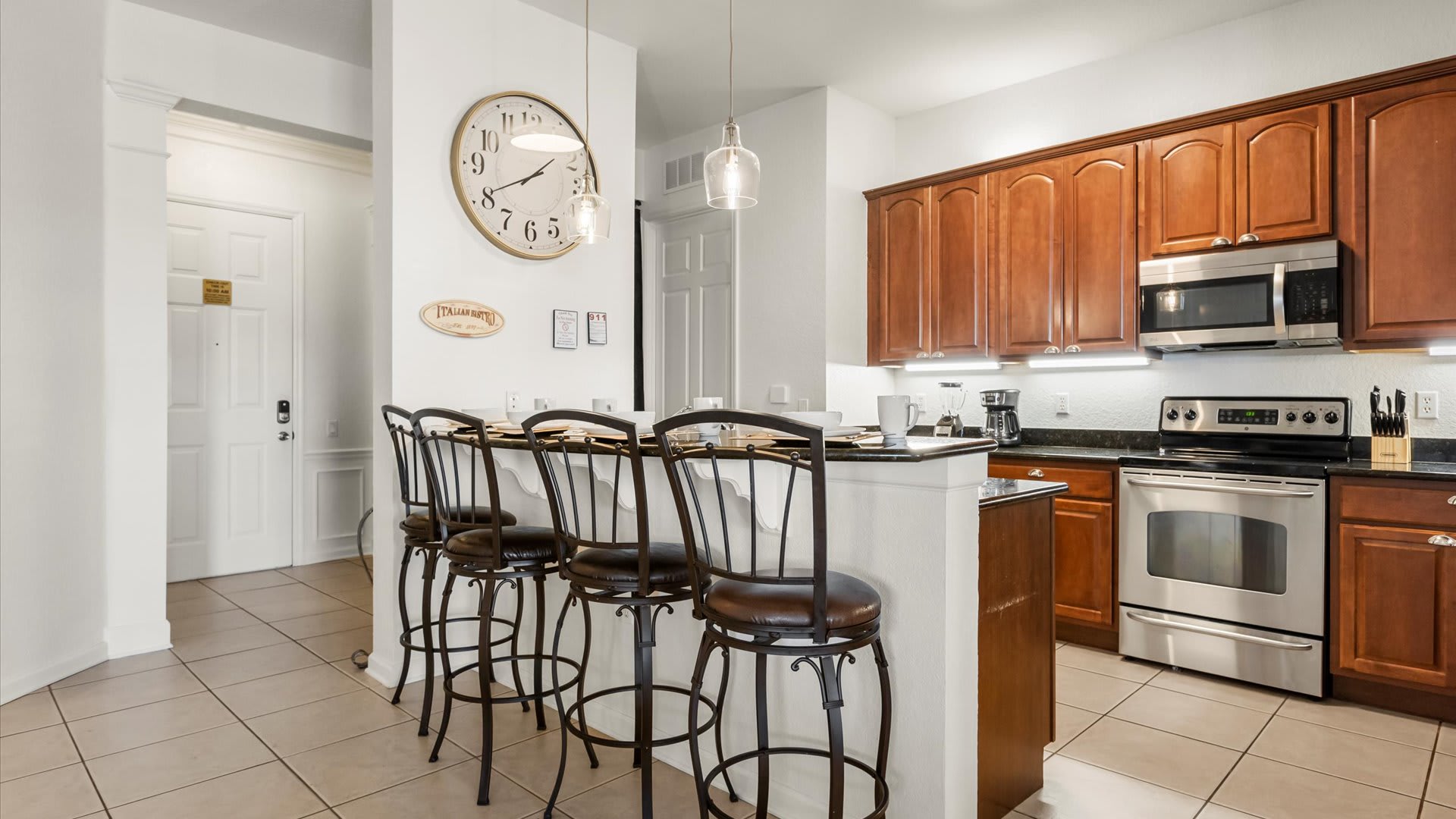 A detailed view of the kitchen's breakfast bar, featuring black granite countertops and four black metal bar stools. A large round wall clock with Roman numerals is a prominent decorative element, with pendant lights illuminating the space.
