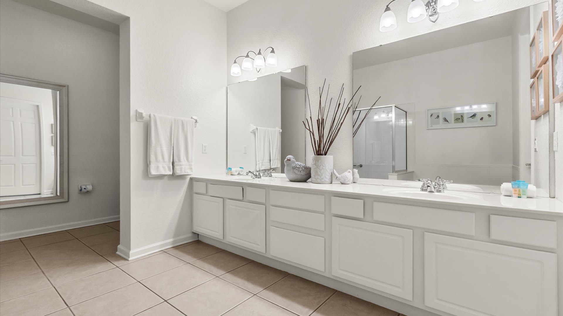 Wide view of bathroom-1, highlighting the extensive white double vanity with ample counter space and two large mirrors, complemented by light tile flooring and white walls. Decorative elements include a vase with branches and bird figurines.