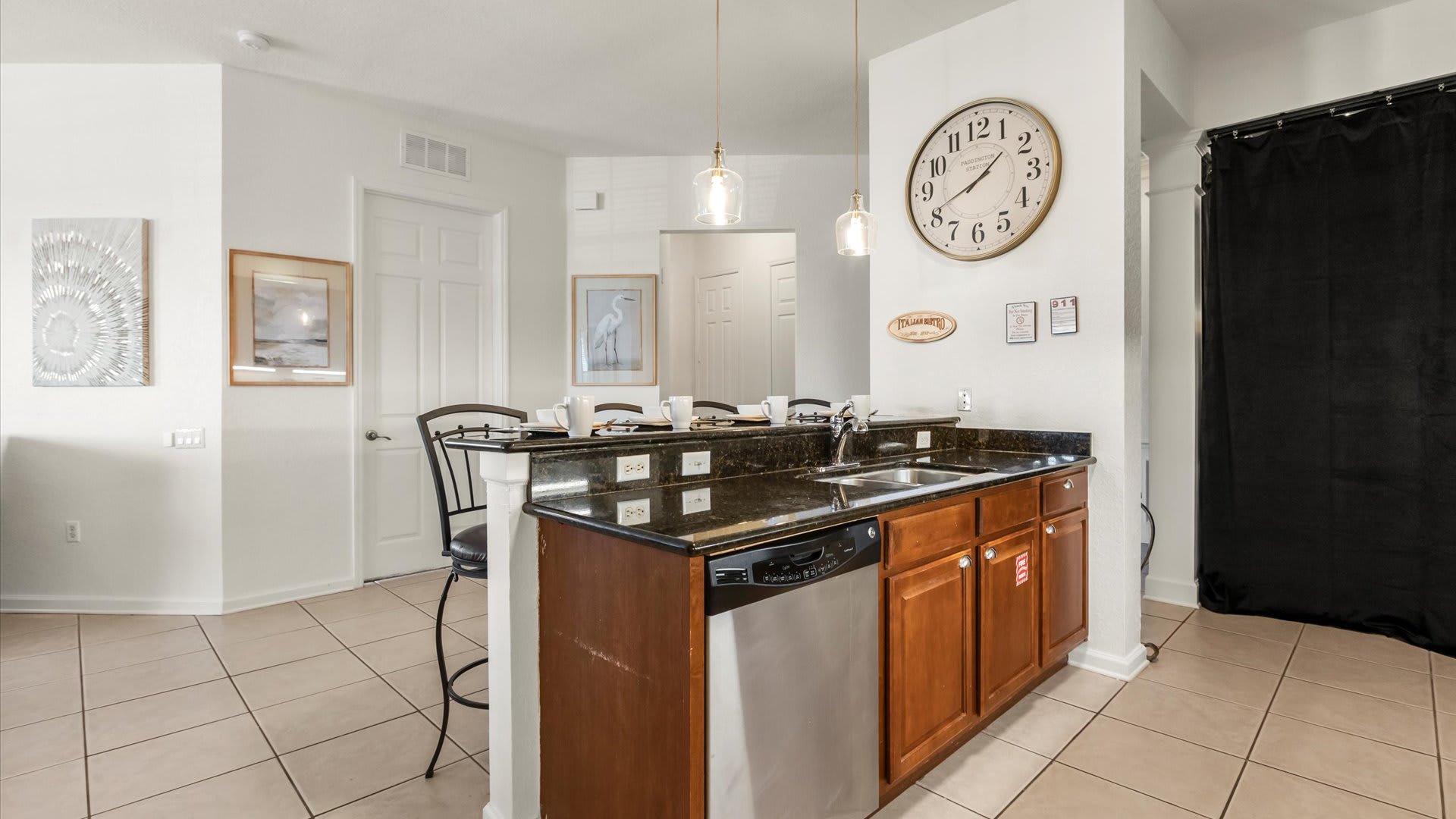 The kitchen's breakfast bar includes a double sink and a stainless steel dishwasher, set within black granite countertops. Wooden cabinets are visible below, and a large wall clock hangs above the bar.
