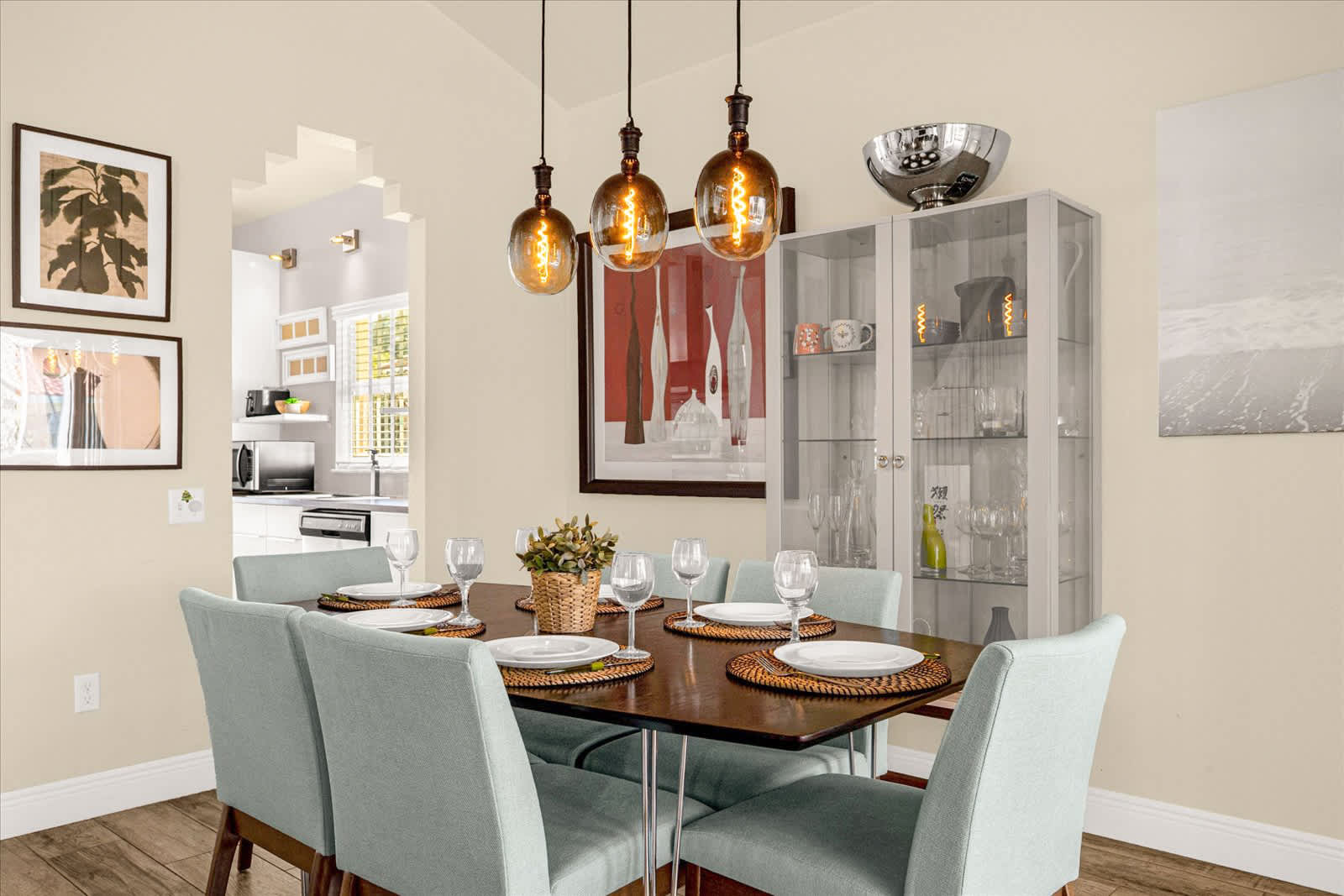 Elegant dining room with a dark wood table set for six, surrounded by light blue upholstered chairs, illuminated by three industrial-style pendant lights, and featuring a glass display cabinet and modern artwork.