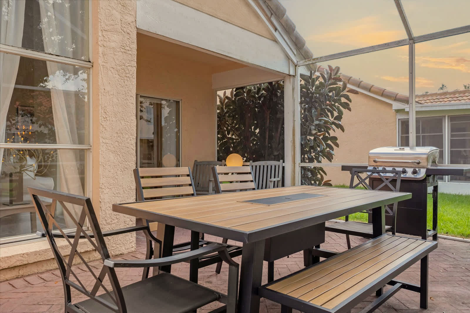 A spacious screened patio features a modern outdoor dining table with a built-in fire pit, surrounded by black metal chairs and a bench, all set on red brick pavers. A gas grill is visible in the background, and the area is covered by a roof extension.