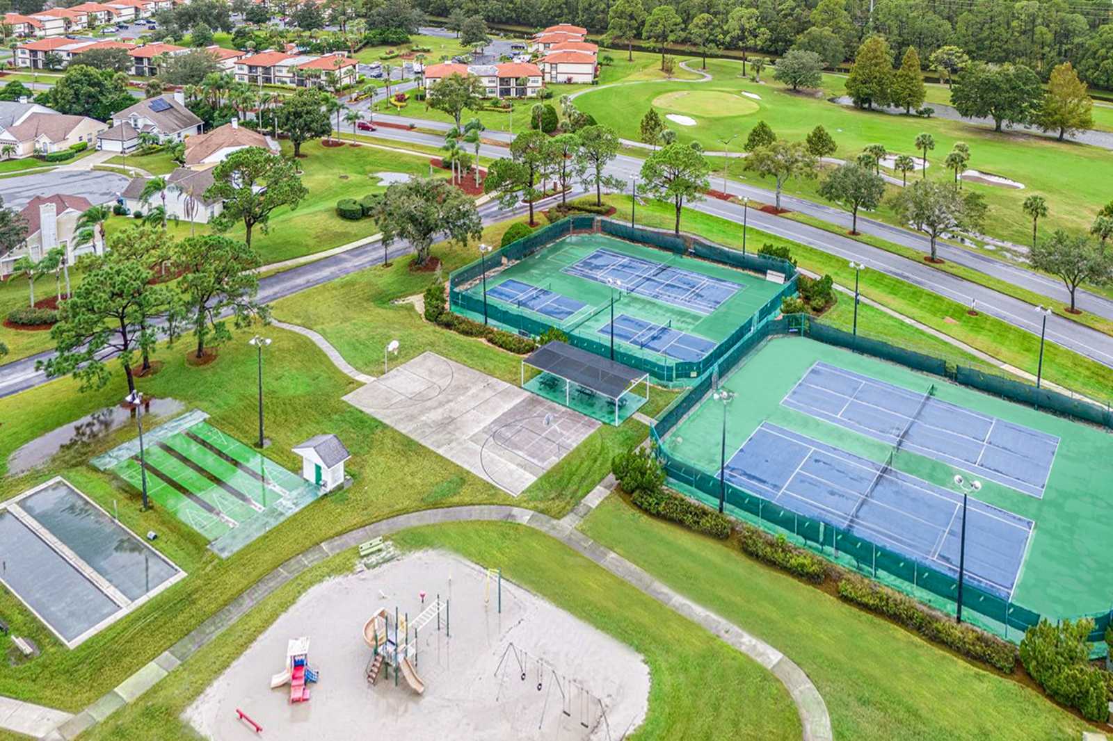 Aerial view of a community recreational area featuring multiple tennis courts, a basketball court, and a children's playground with swings and slides. The facilities are surrounded by expansive green lawns and mature trees, with residential areas and roads visible in the background.
